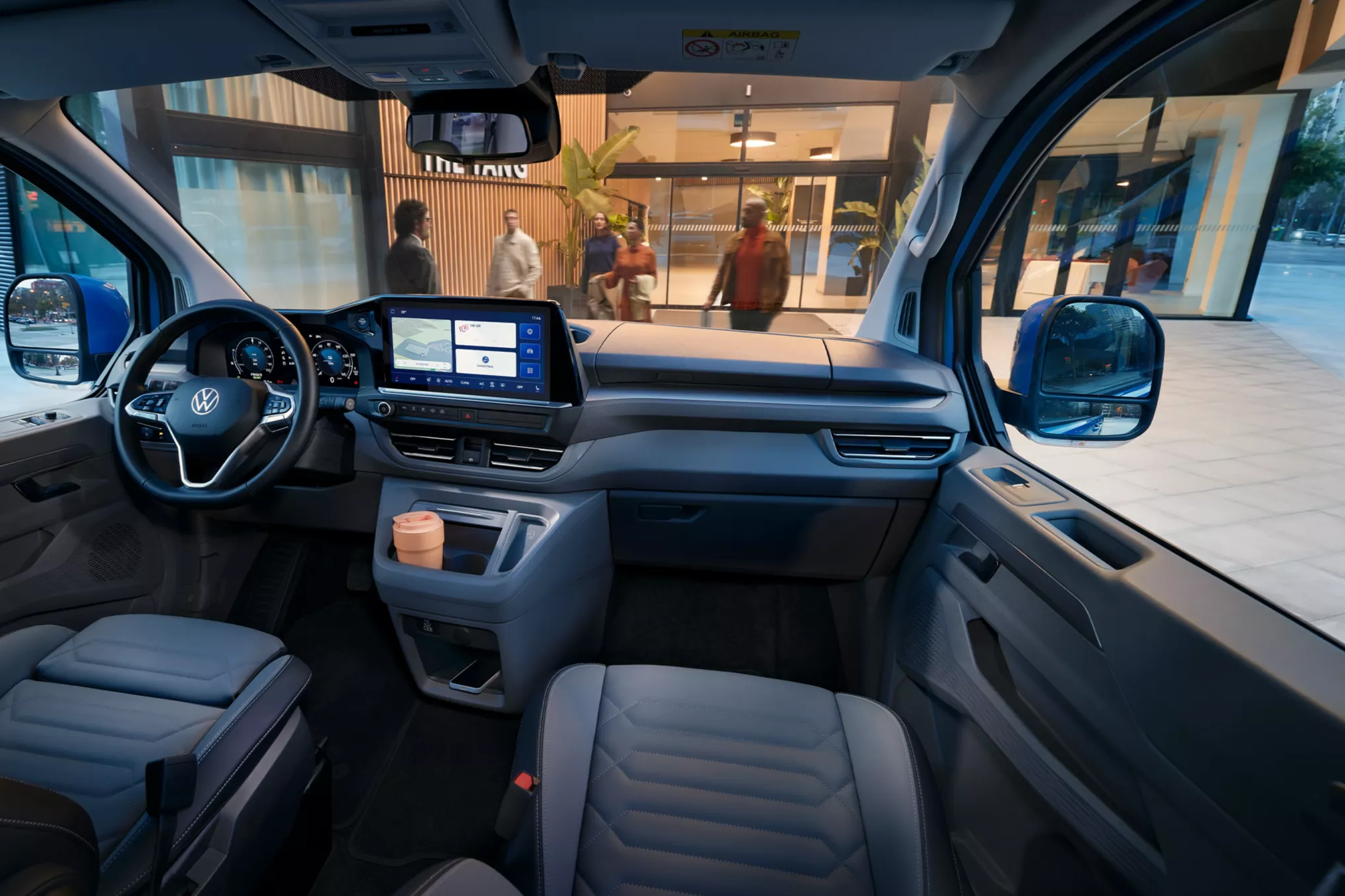 Notranjost sodobnega kombija z zaslonom na dotik na armaturni plošči, prostornimi usnjenimi sedeži in držalom za skodelico ob prestavni ročici. V ozadju je vidno okno, ki razkriva pešce in vhod v stavbo.