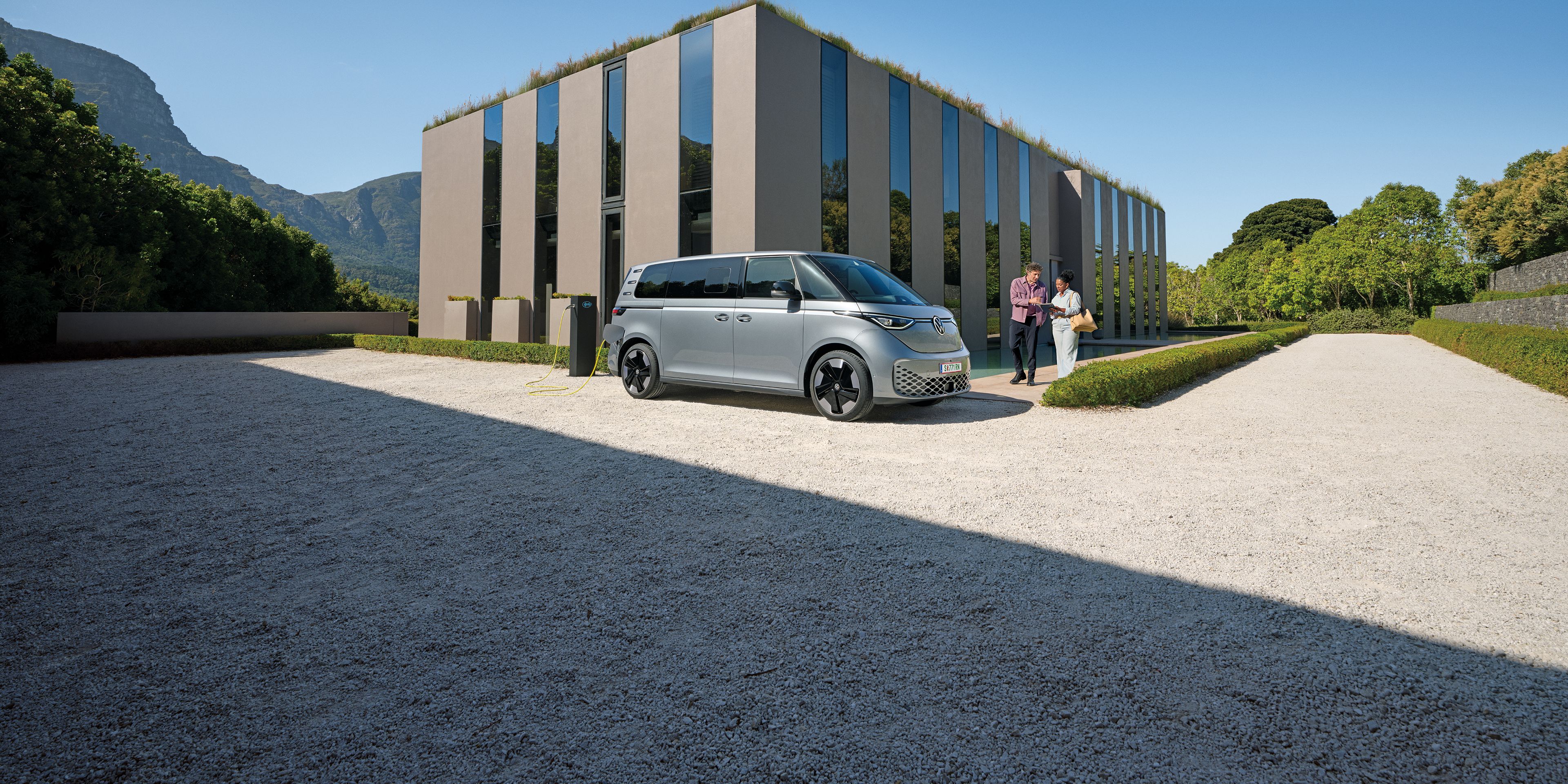 Ein silberner ID. Buzz parkt vor einem modernen Haus, ein Paar  geht auf das Auto zu