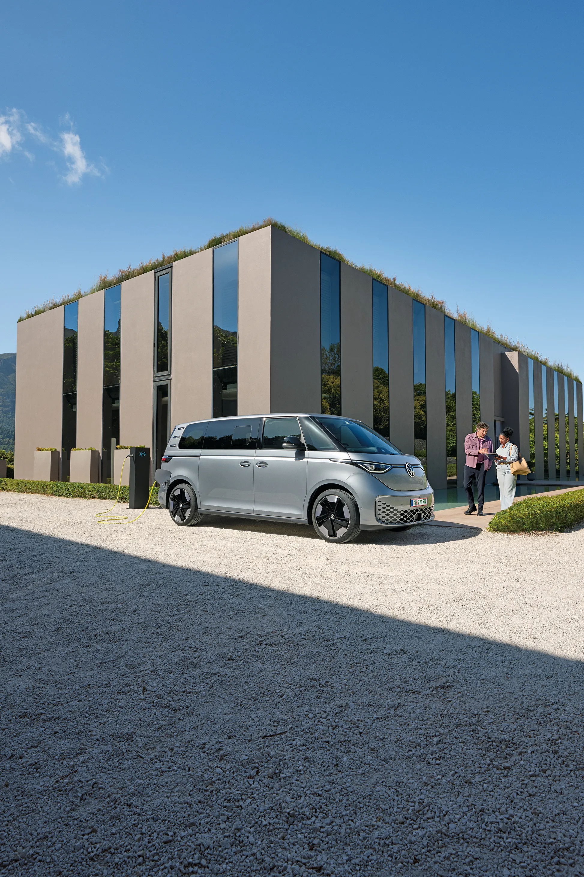 Ein silberner ID. Buzz parkt vor einem modernen Haus, ein Paar  geht auf das Auto zu