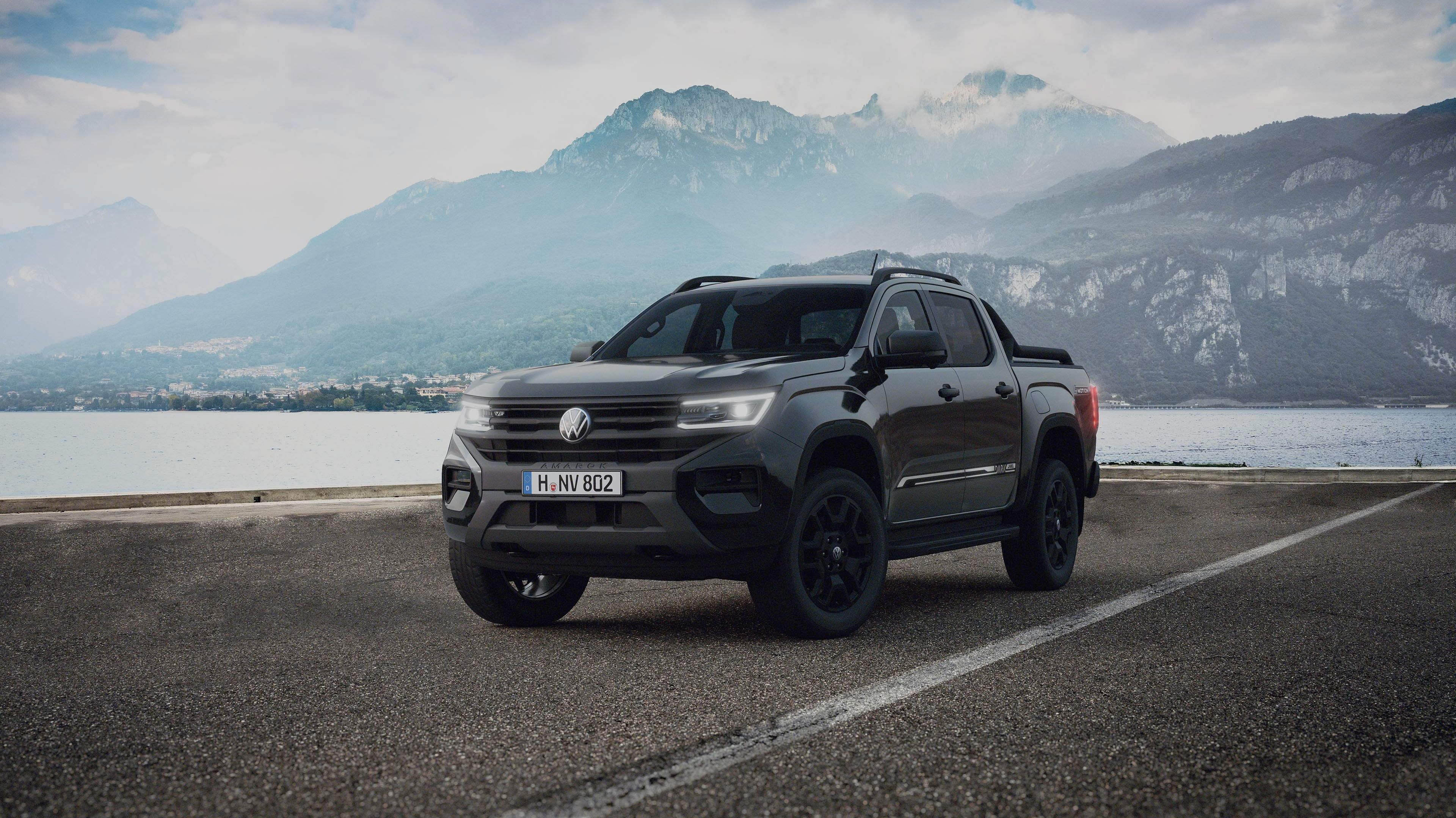 Temno siv pick-up stoji na asfaltirani cesti ob jezeru, v ozadju pa se dvigajo megleno modre gorske verige. Nebo je delno oblačno, kar ustvarja dramatično vzdušje. Avto je obrnjen rahlo v levo, poudarjeni so njegovi robustni in športni detajli.