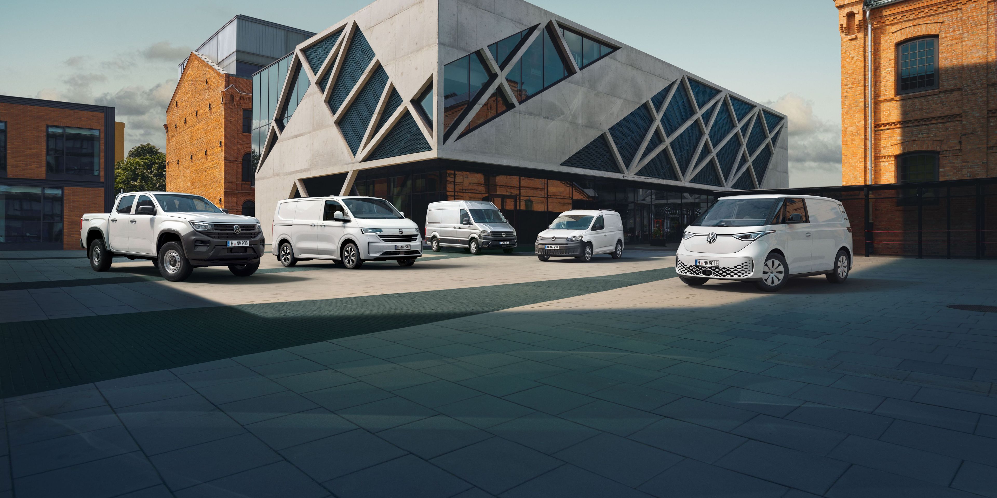 Pred stavbo z modernimi arhitekturnimi elementi je parkiranih več belih dostavnih vozil. Stavba ima velike steklene površine in zanimive geometrijske oblike. V ozadju so vidne starejše zgradbe iz opeke.