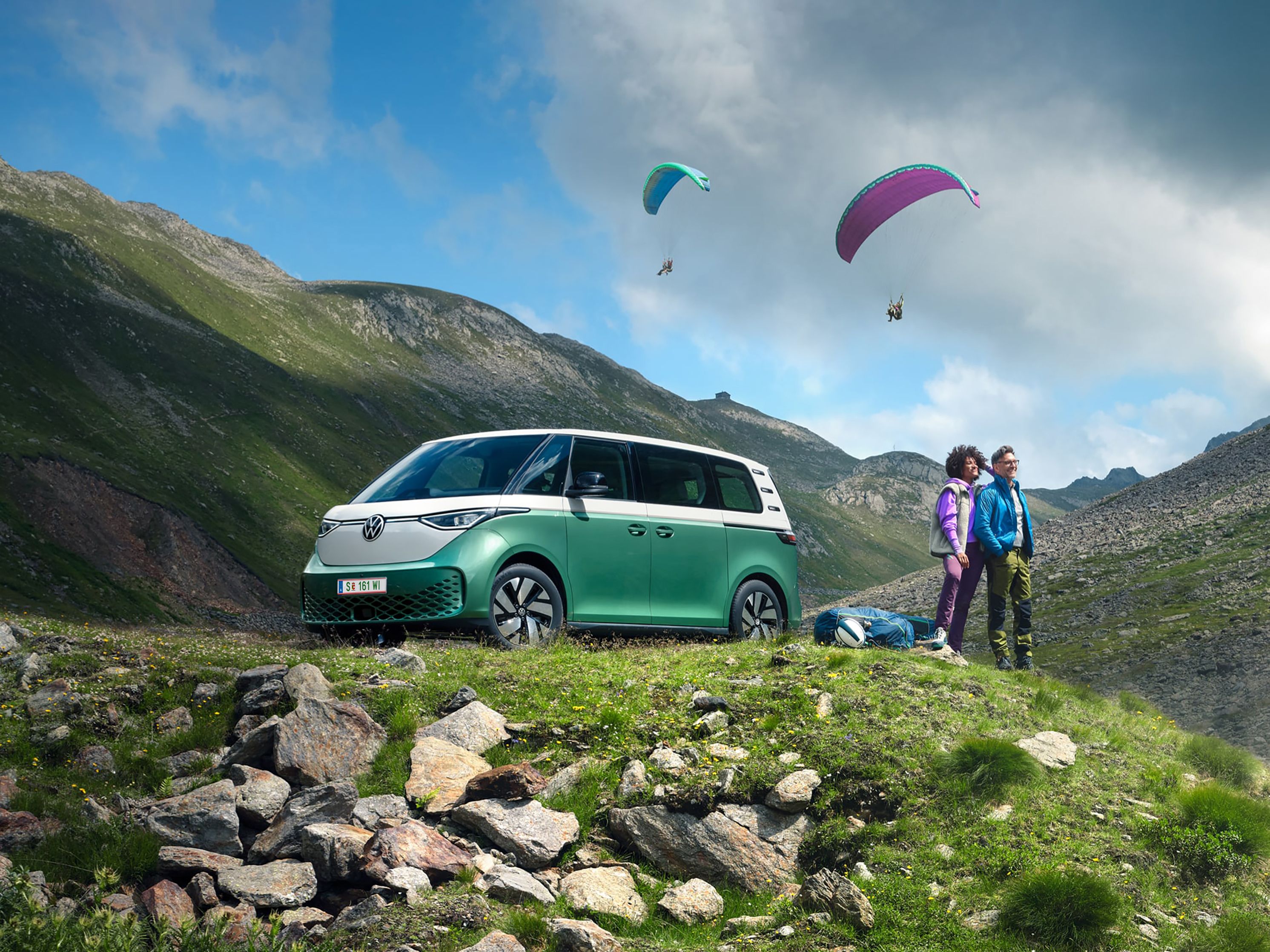 Ein grüner Volkswagen ID. Buzz parkt an einer Bergstraße, ein Mann und eine Frau beobachten Paragleiter.