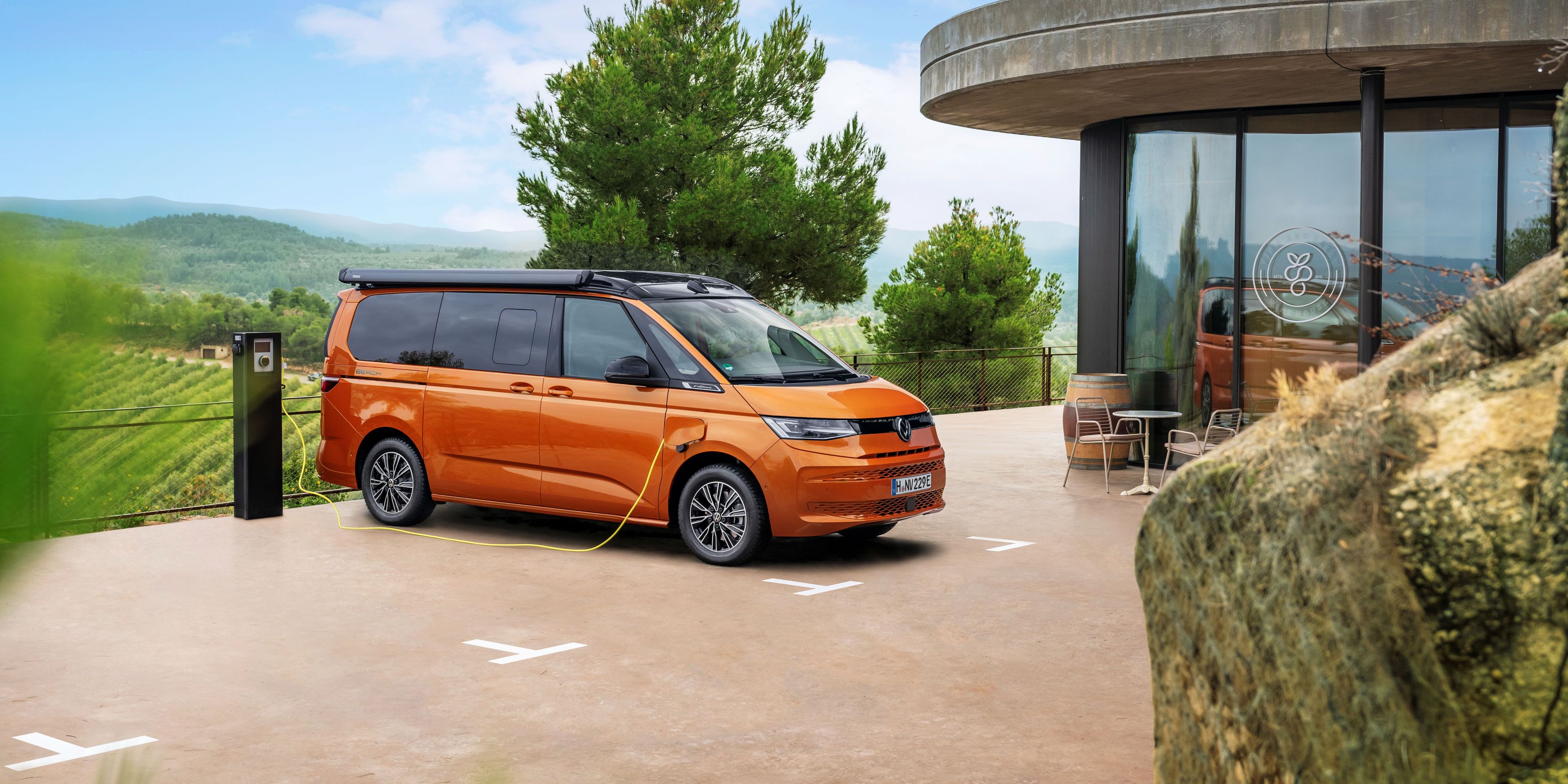 Oranžen kombi je parkiran ob sodobni zgradbi na polnilni postaji za električna vozila, obdan z zelenimi polji in drevesi. Zgradba ima steklene stene in zunanjo teraso s stoli in mizami. V ozadju se razprostirajo griči pod modrim nebom z nekaj oblaki.