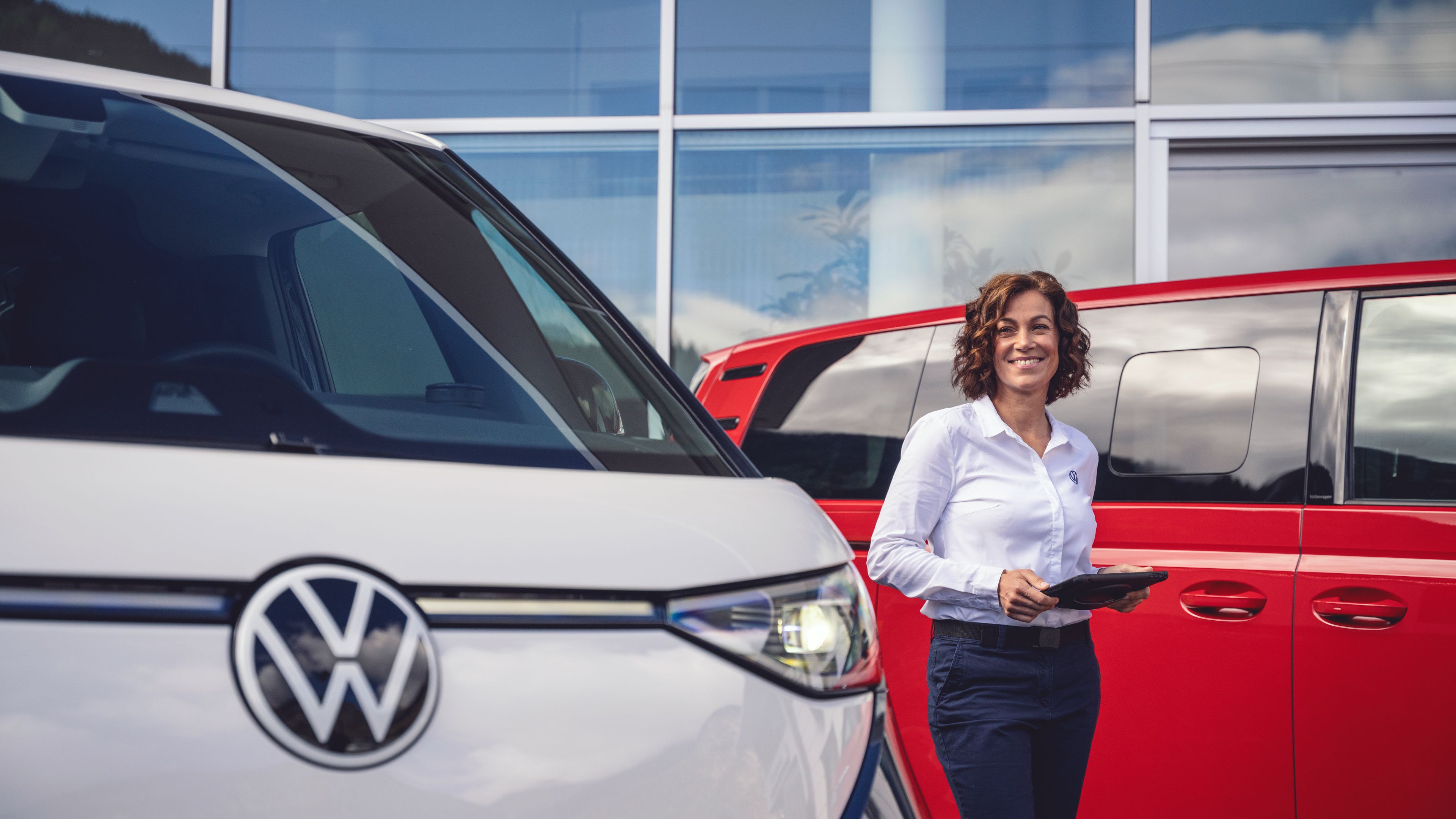 Nasmejana prodajalka avtomobilov stoji pred sodobnim prodajnim salonom, v rokah drži tablico. Ob njej sta parkirana bela in rdeča kombinirana vozila znamke Volkswagen. Ozadje tvorijo steklena fasada stavbe in odsevajoče nebo z oblaki.