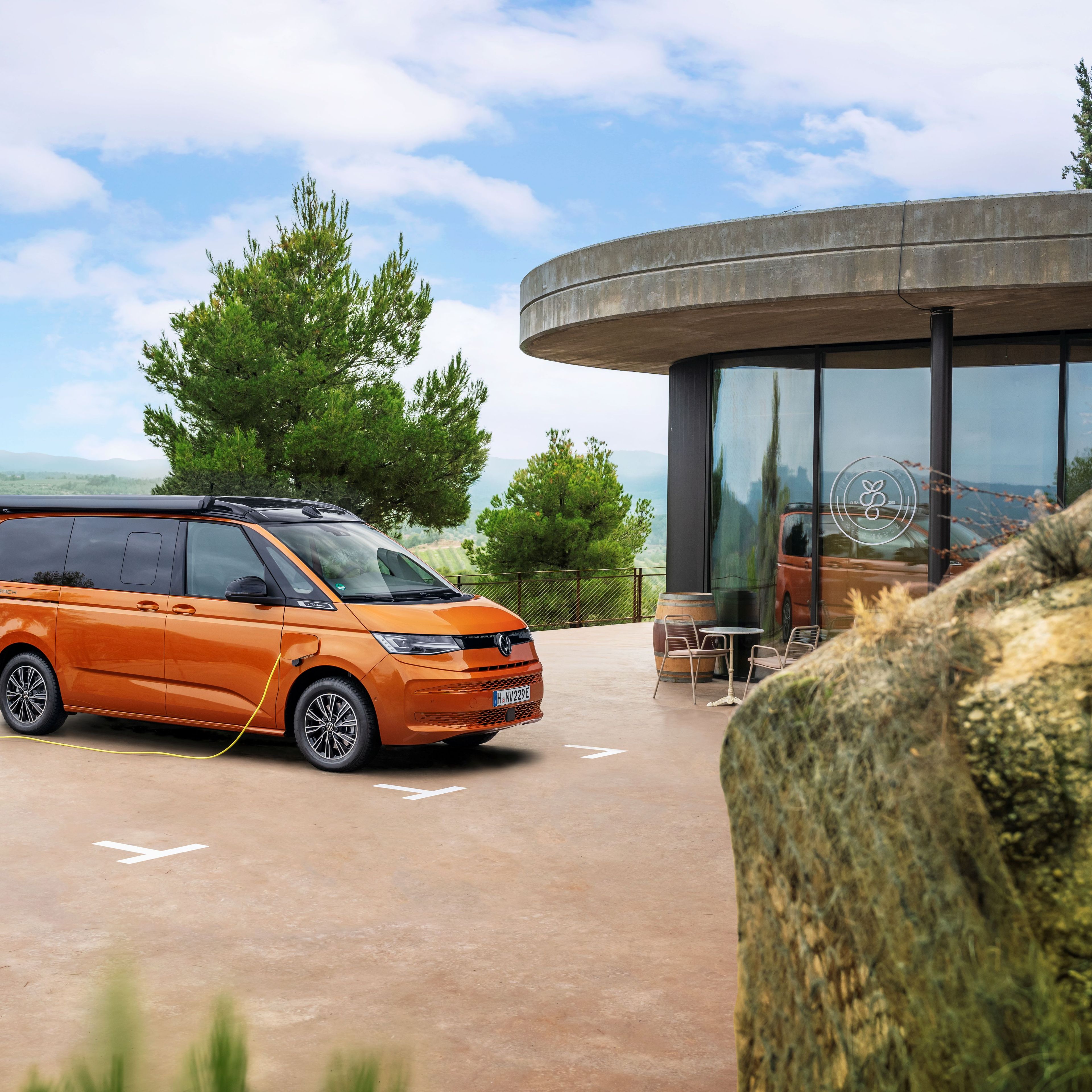 Oranžen kombi je parkiran ob sodobni zgradbi na polnilni postaji za električna vozila, obdan z zelenimi polji in drevesi. Zgradba ima steklene stene in zunanjo teraso s stoli in mizami. V ozadju se razprostirajo griči pod modrim nebom z nekaj oblaki.