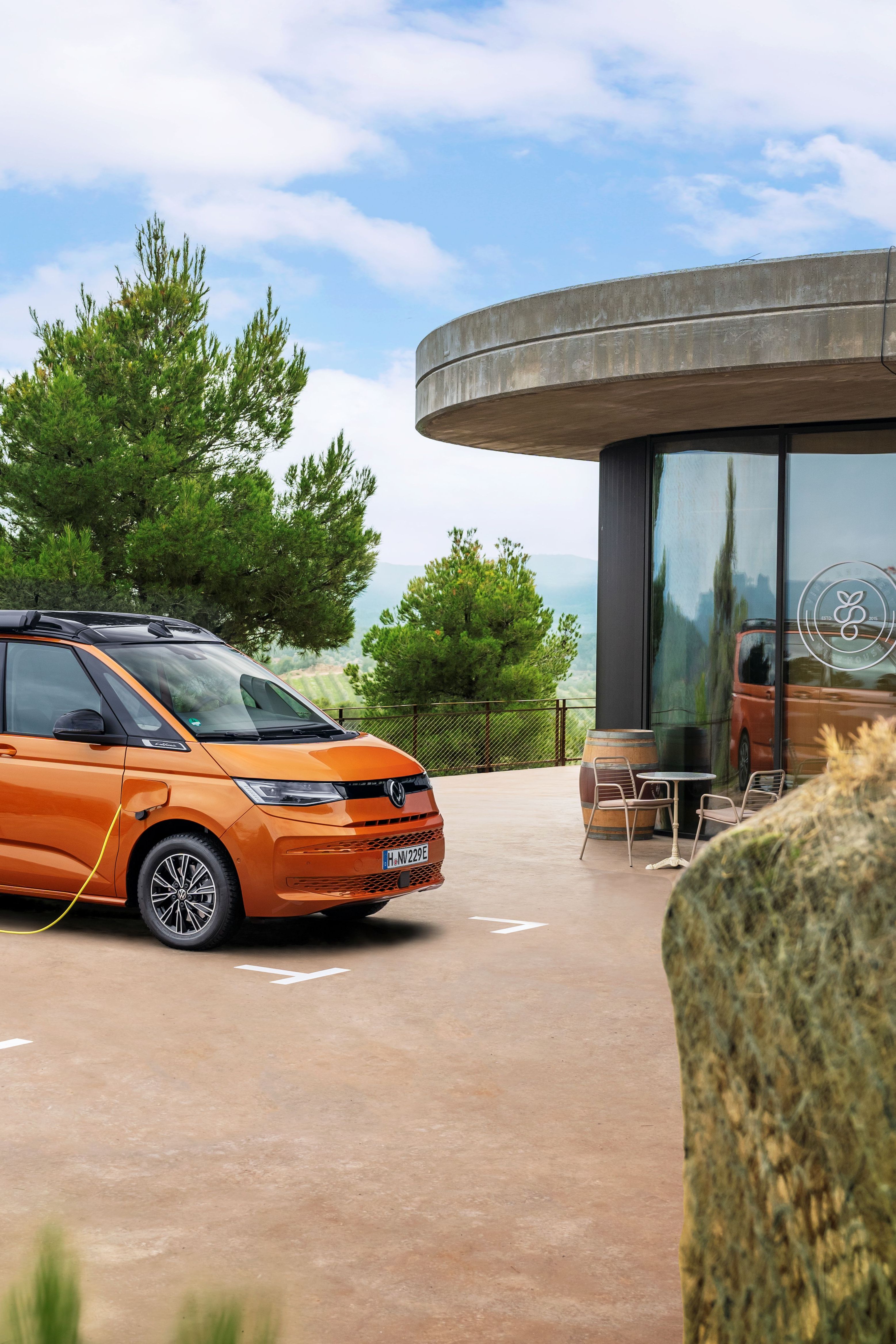 Oranžen kombi je parkiran ob sodobni zgradbi na polnilni postaji za električna vozila, obdan z zelenimi polji in drevesi. Zgradba ima steklene stene in zunanjo teraso s stoli in mizami. V ozadju se razprostirajo griči pod modrim nebom z nekaj oblaki.