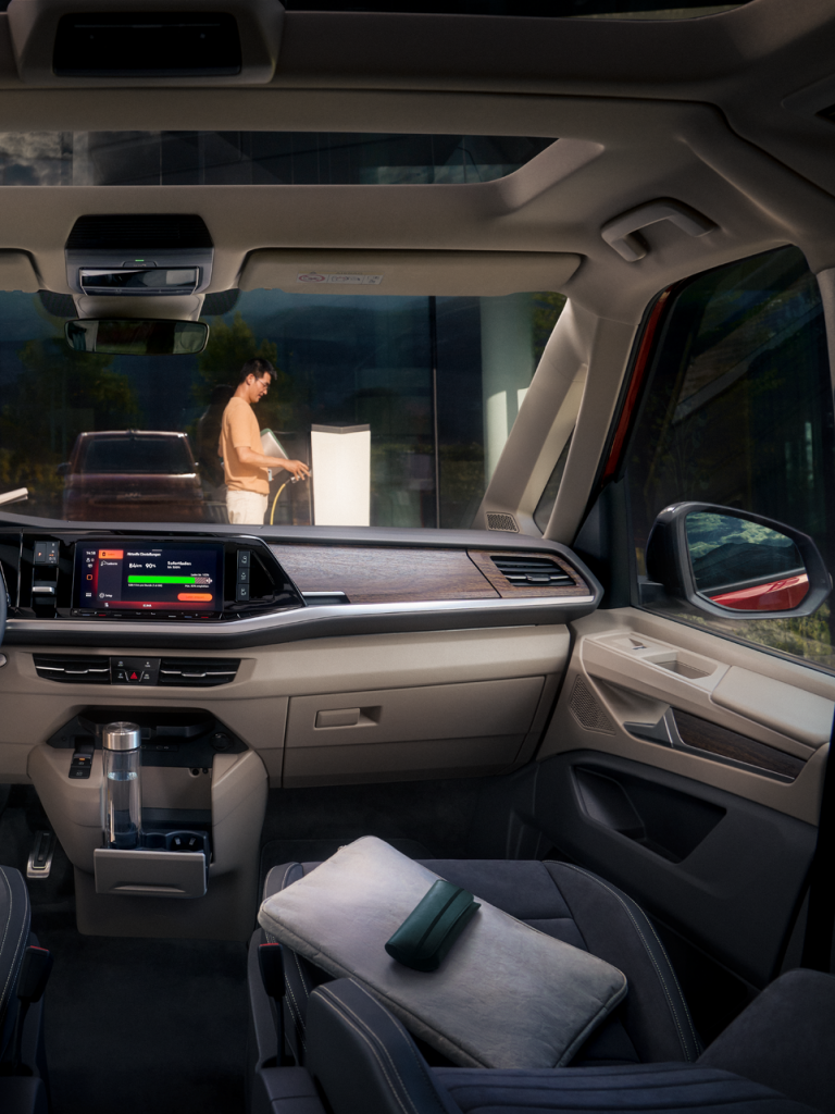 Notranjost sodobnega avtomobila s širokim vetrobranskim steklom. Osrednji del armaturne plošče ima velik zaslon na dotik. Dva človeka stojita zunaj avtomobila ob steklu.