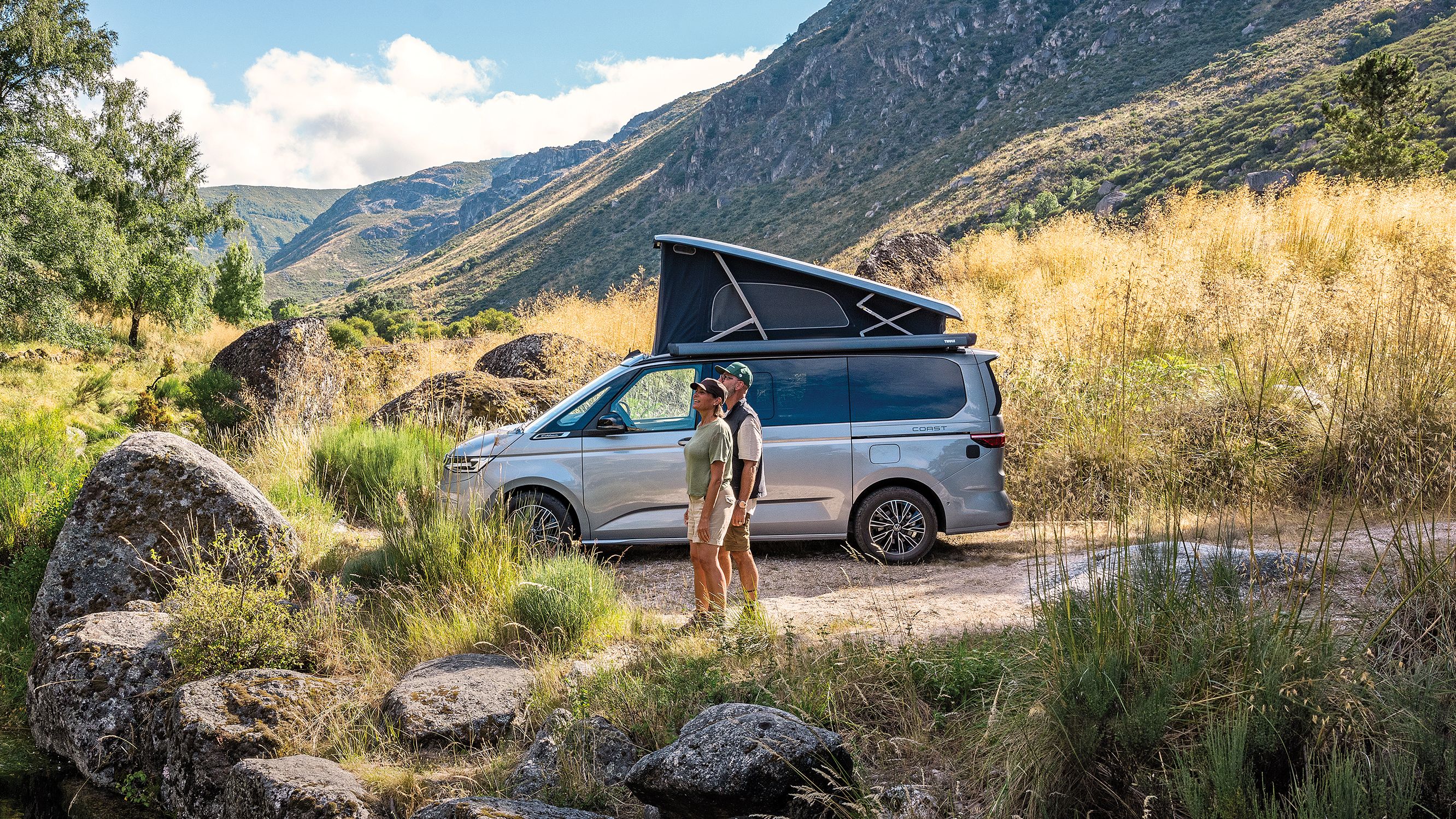 Ein New California mit ausgeklapptem Dach parkt an einem See, ein Paar steht am Seeufer