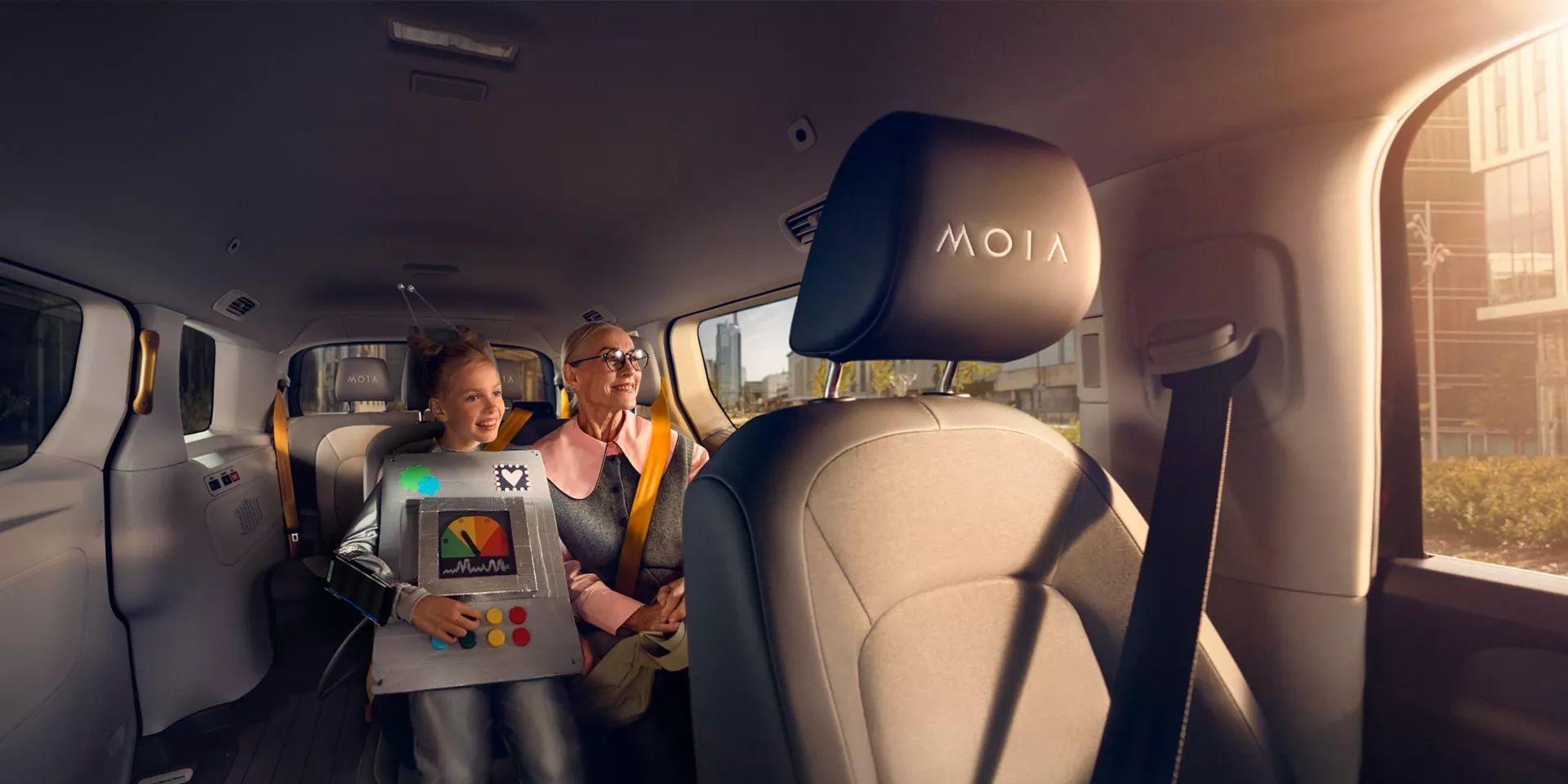 Ein Kind sitzt auf dem Rücksitz eines Autos und spielt auf einem Tablet, während ein Erwachsener neben ihm sitzt. Die Sitze haben einen schwarzen Bezug mit dem Logo "MOIA". Das Auto ist geräumig und von Tageslicht durchflutet.