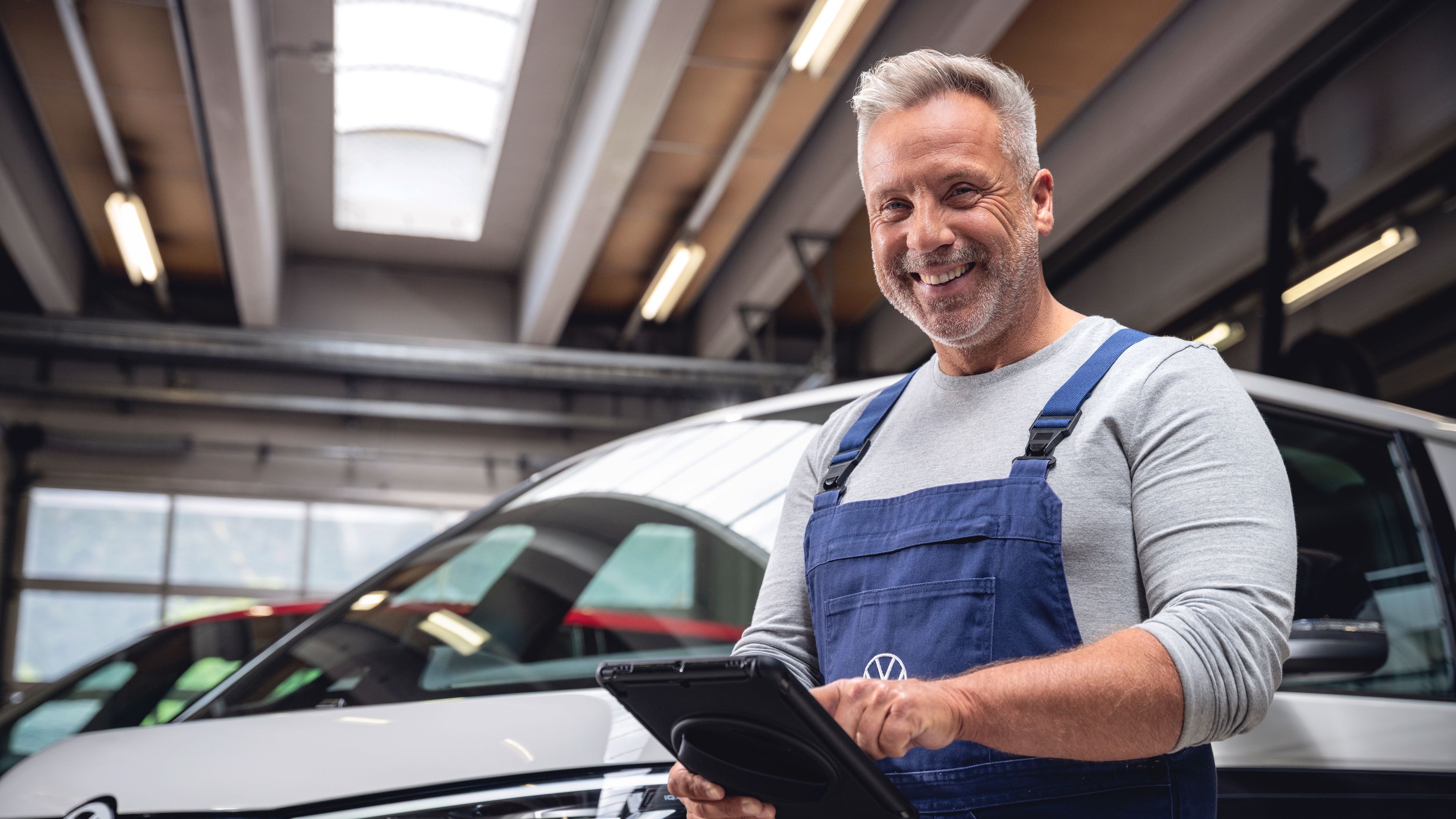 Sivolasi avtomehanik v modrem delovnem kombinezonu stoji v delavnici in se nasmiha, medtem ko v rokah drži tablico. Za njim je parkirano belo vozilo znamke Volkswagen, delno vidno v prostoru z industrijsko razsvetljavo. Atmosfera je profesionalna in prijazna, kot pri servisnem pregledu vozila.