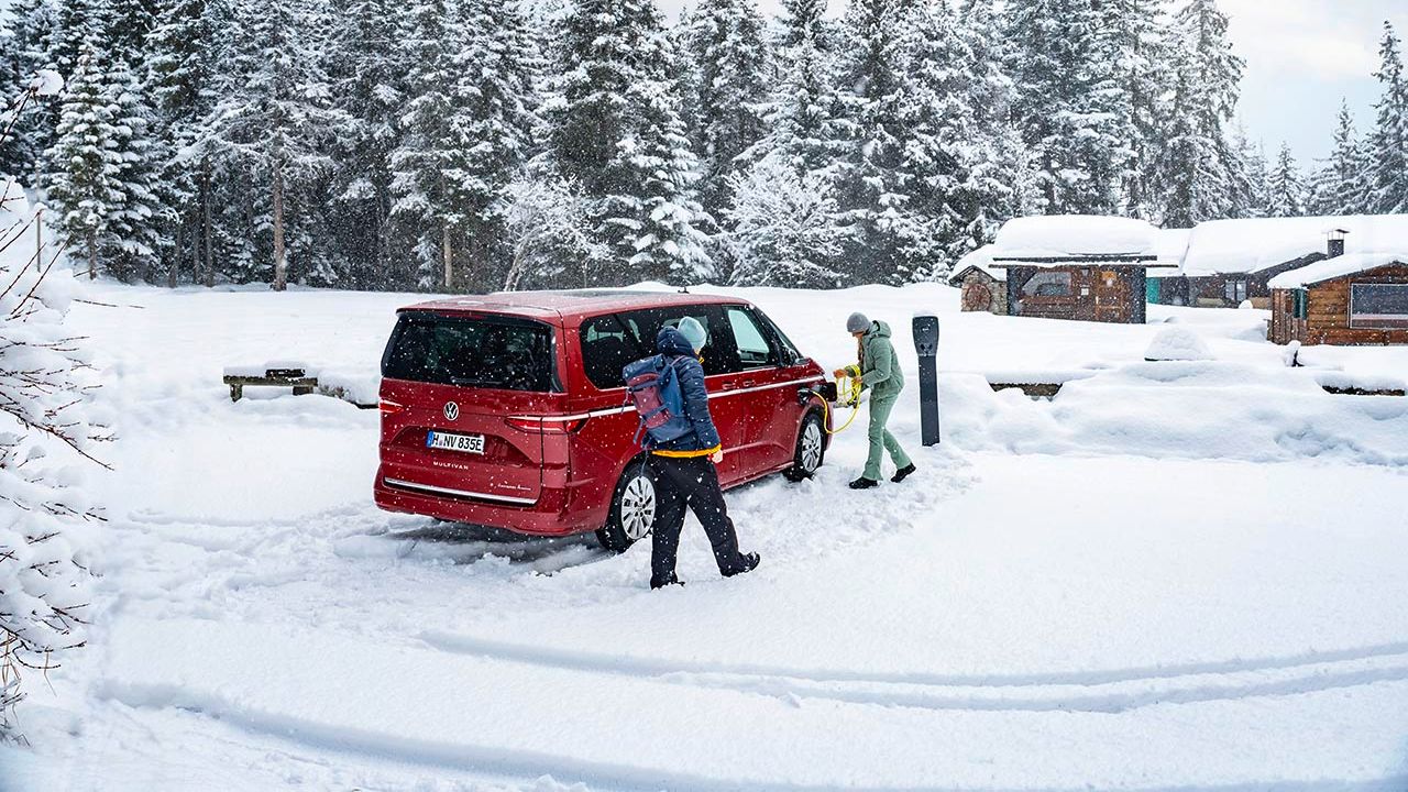 Dva človeka v zimski obleki stojita ob rdečem kombiju na snegu, ki je parkiran ob polnilni postaji. V ozadju je gozd in nekaj lesenih koč, vse prekrito s snegom. Nebo je oblačno.