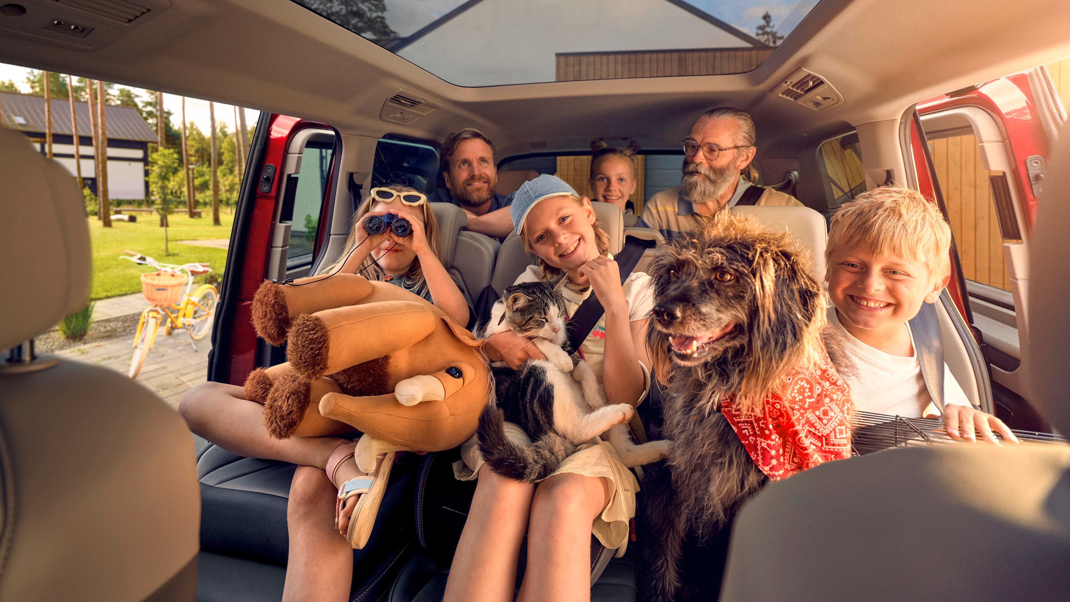 Skupina ljudi, ki sedijo v avtomobilu, nekdo drži plišaste igrače, drugi osebi pa sedi v naročju pes in mačka. Avto ima panoramsko streho, zunaj pa je sončen dan. V ozadju je vidna hiša in kolo.