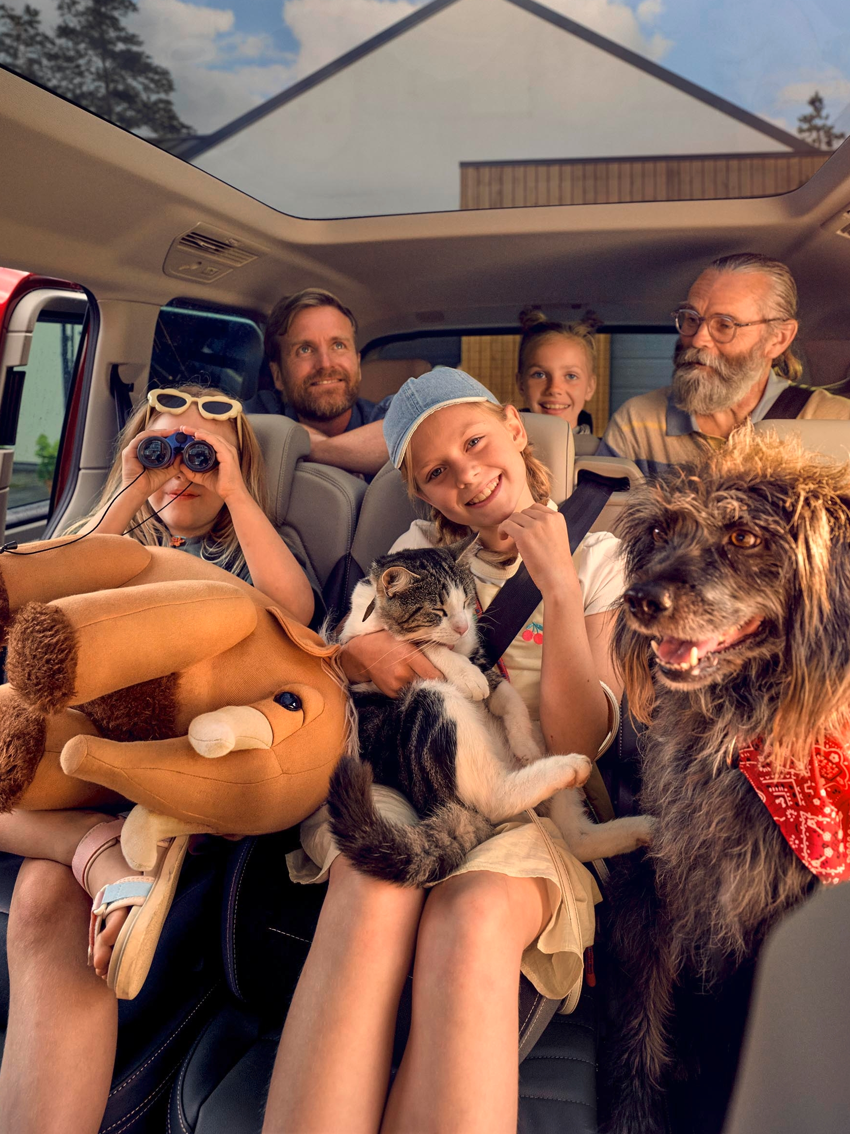 Skupina ljudi, ki sedijo v avtomobilu, nekdo drži plišaste igrače, drugi osebi pa sedi v naročju pes in mačka. Avto ima panoramsko streho, zunaj pa je sončen dan. V ozadju je vidna hiša in kolo.