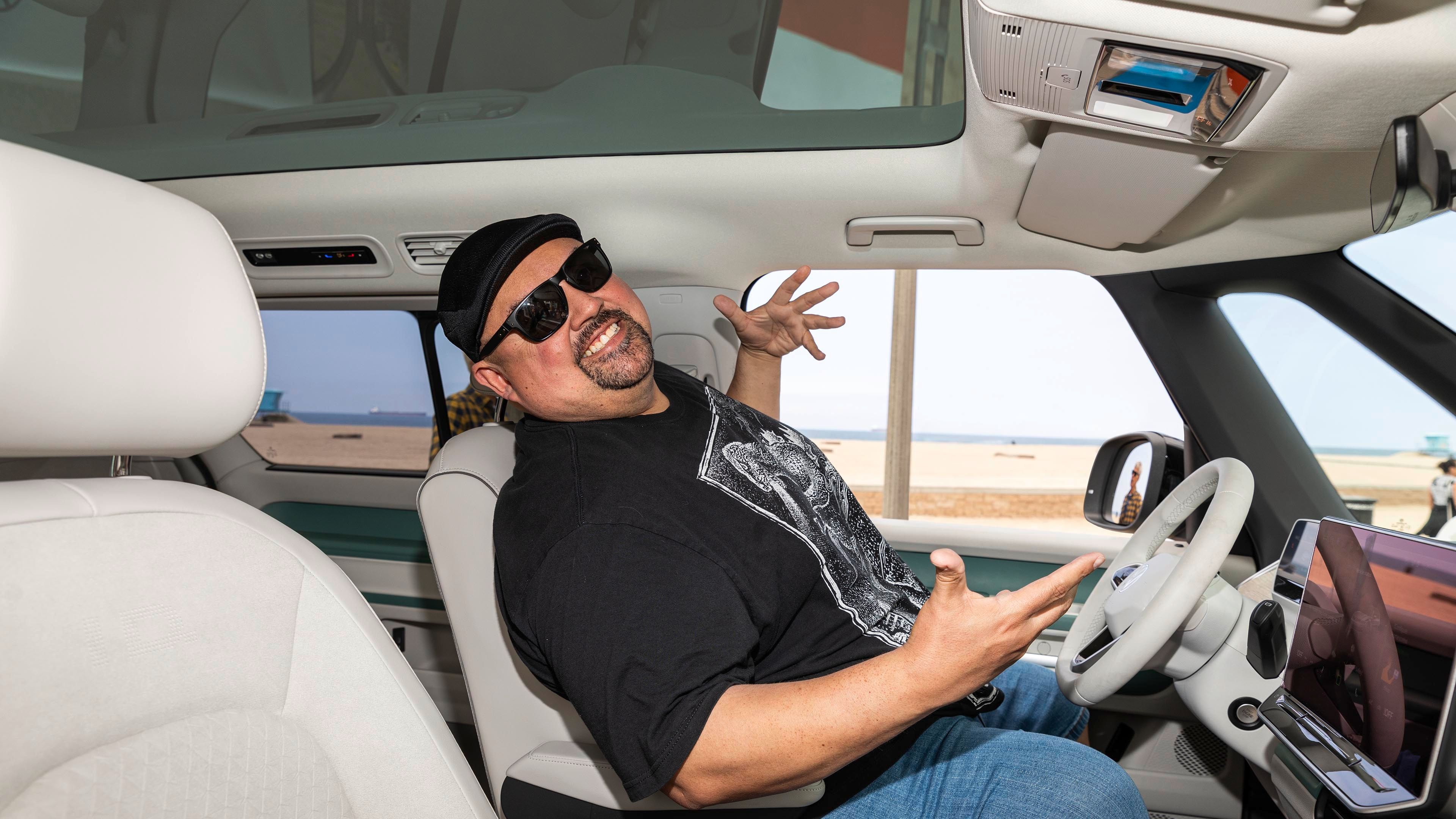 Oseba sedi na voznikovem sedežu v avtomobilu ob morju in živahno gestikulira z rokami. Odprta vrata omogočajo pogled na peščeno plažo v ozadju.