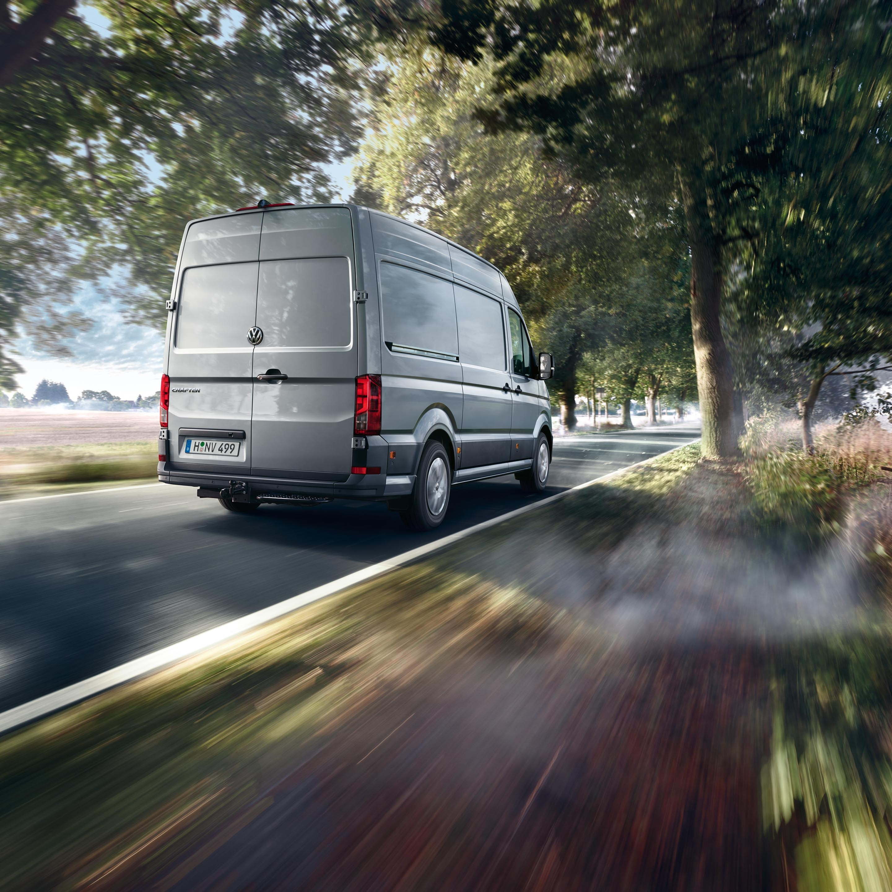 Siva dostavna kombi vozi po podeželski cesti, obdani z drevesi. Zgodaj zjutraj meglica prekriva del ceste. Pokrajina v ozadju je mirna in slikovita.