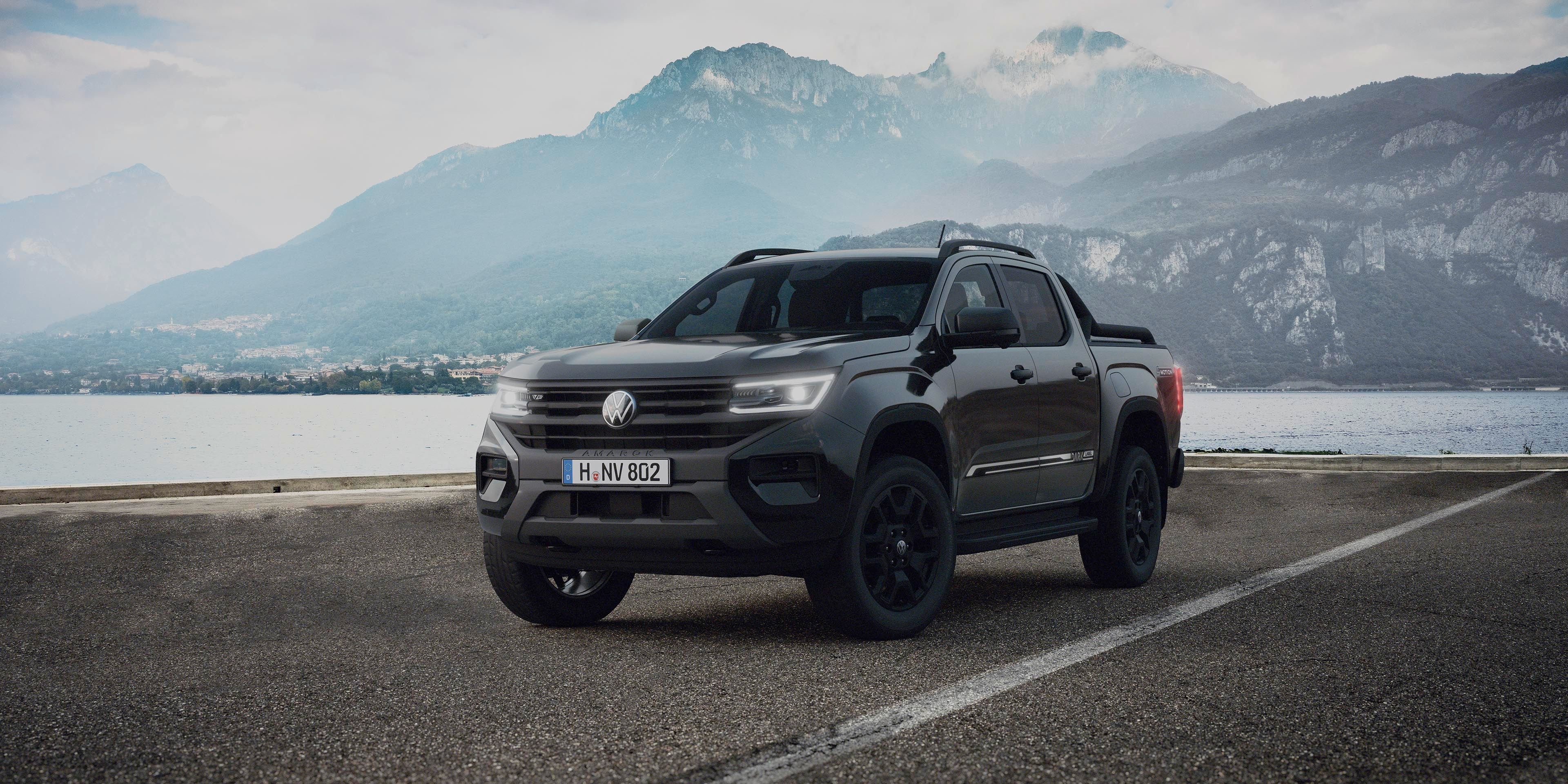 Temno siv pick-up stoji na asfaltirani cesti ob jezeru, v ozadju pa se dvigajo megleno modre gorske verige. Nebo je delno oblačno, kar ustvarja dramatično vzdušje. Avto je obrnjen rahlo v levo, poudarjeni so njegovi robustni in športni detajli.