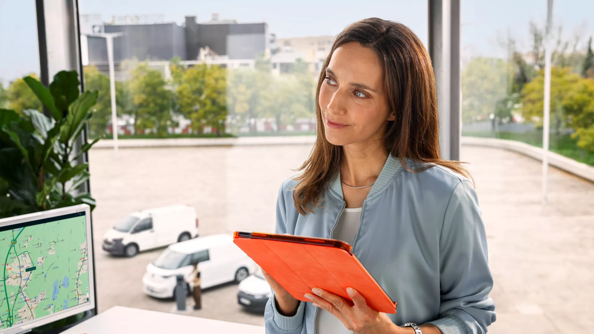 Eine Frau hält ein Tablet in den Händen und steht vor einem großen Fenster mit Blick auf parkende Autos und Bäume. Ein Bildschirm links zeigt eine Karte oder ein Navigationssystem. Die Frau hat eine verschwommene Gesichtspartie.