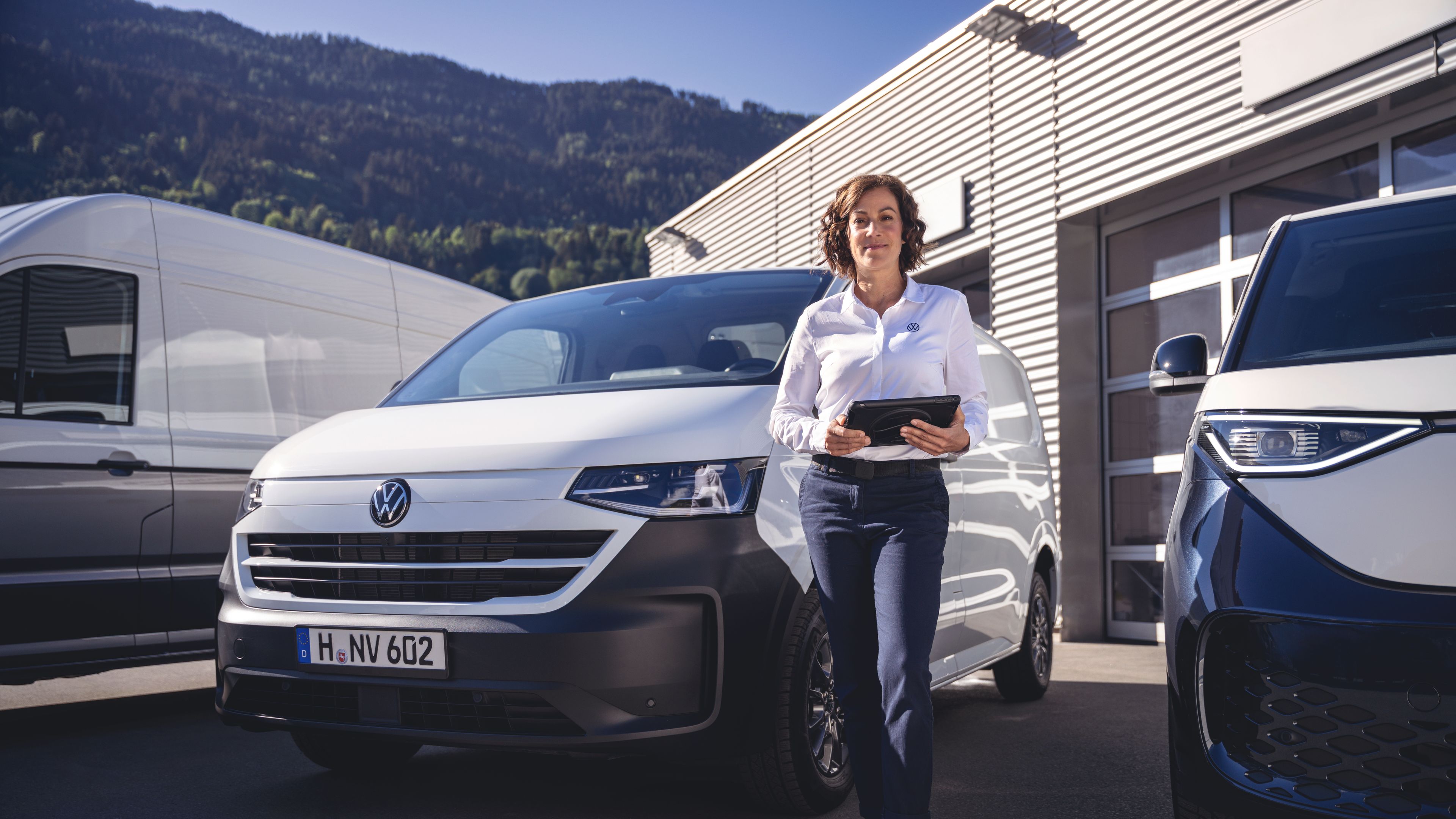 Oseba s tablico v rokah se naslanja na bel kombi znamke Volkswagen, parkiran pred sodobno poslovno stavbo. Ob njej stojita še dva druga dostavna oziroma osebna vozila, v ozadju pa so vidni zeleni hribi ali gore. Podoba deluje kot prizor iz poslovnega ali prodajnega okolja avtomobilov.