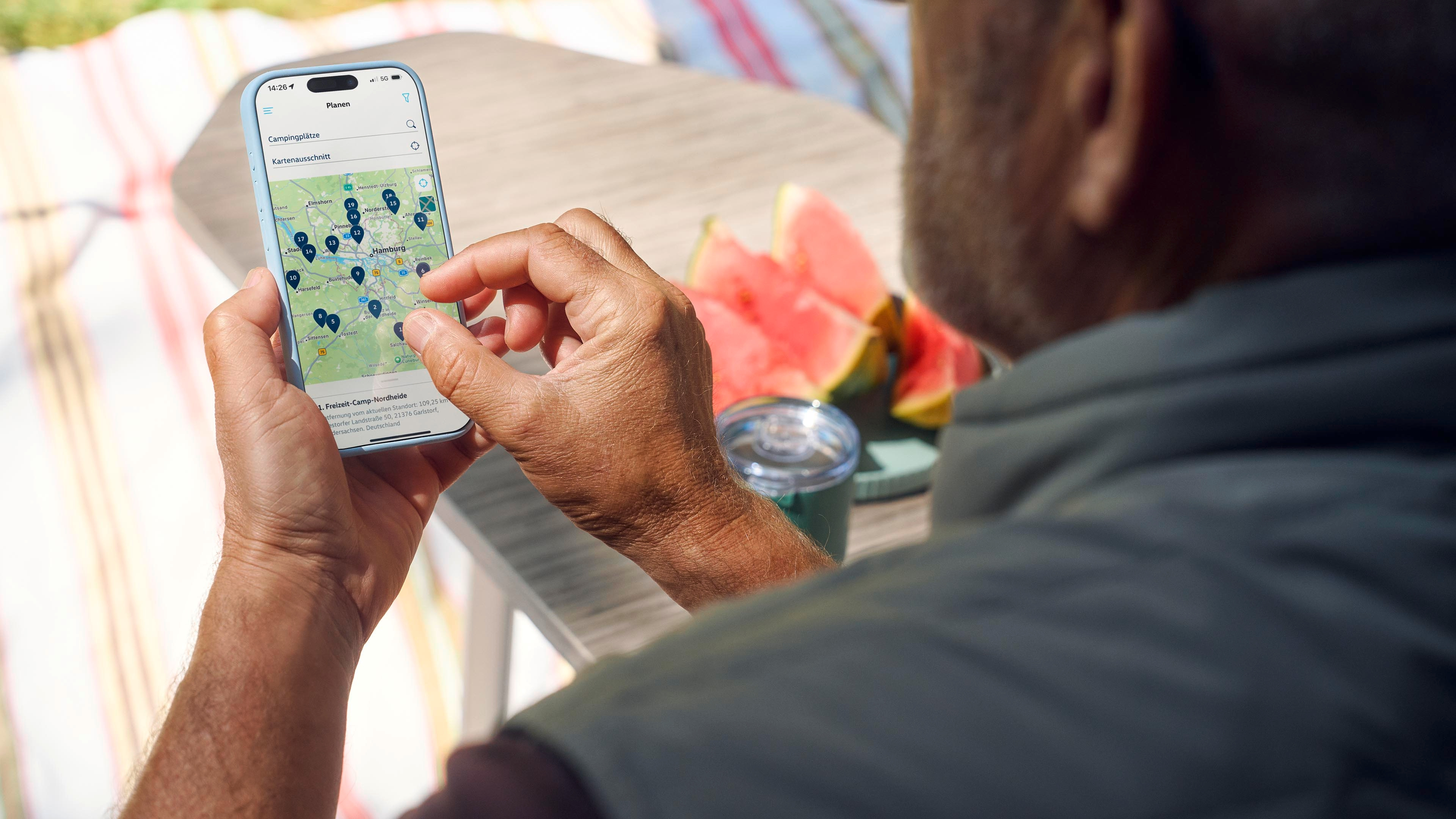 Moški drži mobilni telefon in gleda zemljevid z več modrimi oznakami. Ob njem na mizi sta koščka lubenice in kozarec. V ozadju je odeja položena na travo.