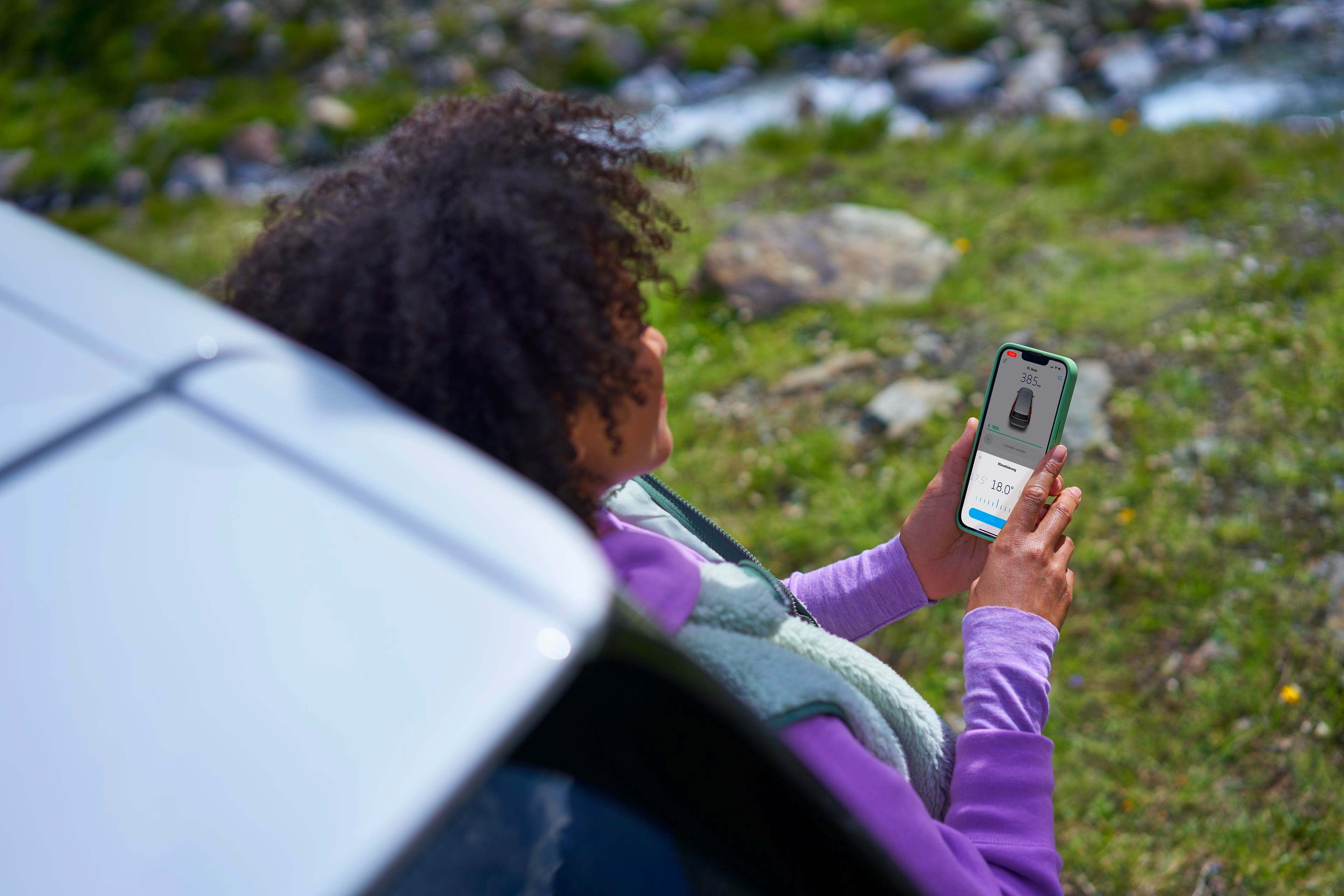 Oseba sedi zunaj poleg avtomobila in s pametnim telefonom preverja informacijo o polnilni postaji za električna vozila. V ozadju je naravna pokrajina s travo, kamni in vodo.