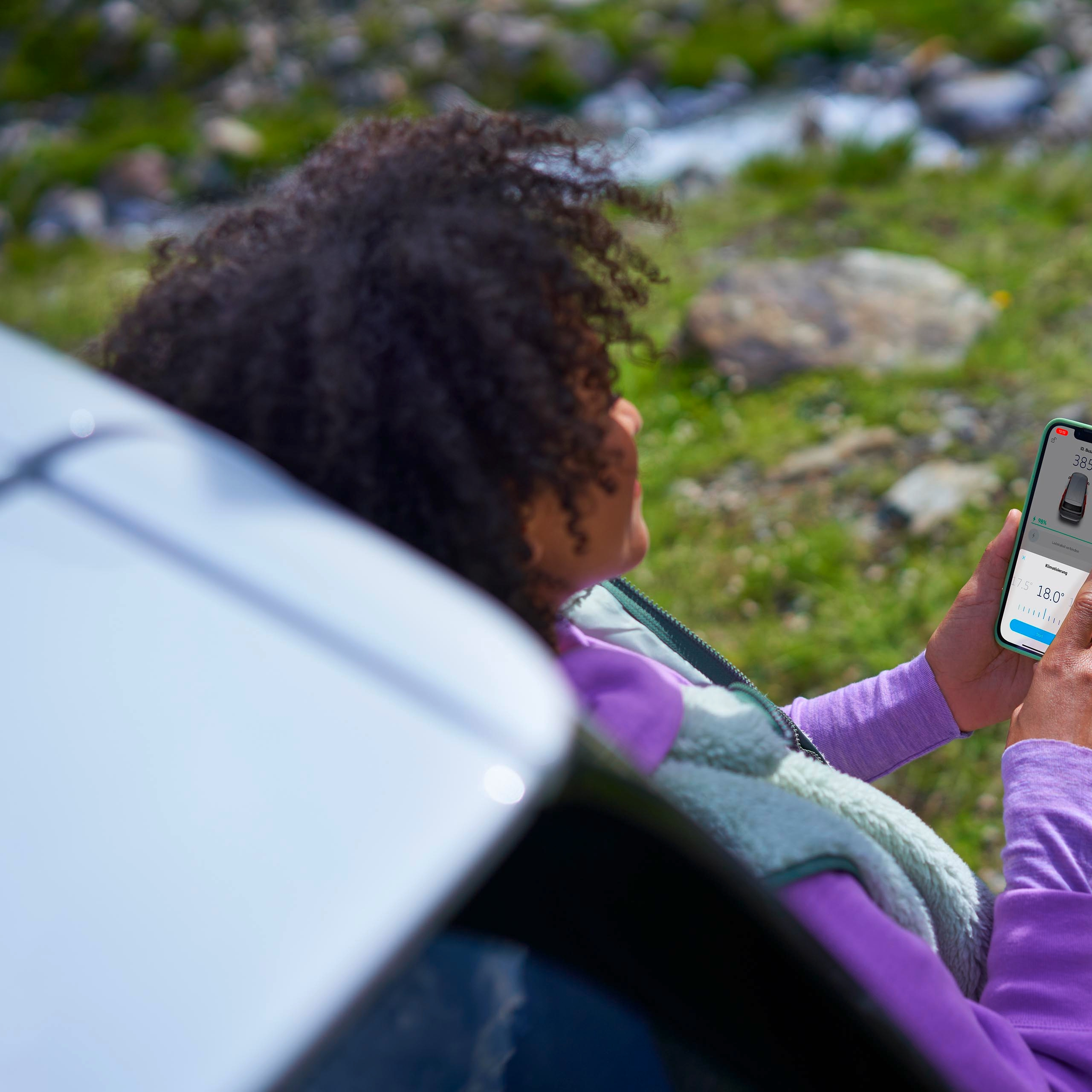Oseba sedi zunaj poleg avtomobila in s pametnim telefonom preverja informacijo o polnilni postaji za električna vozila. V ozadju je naravna pokrajina s travo, kamni in vodo.