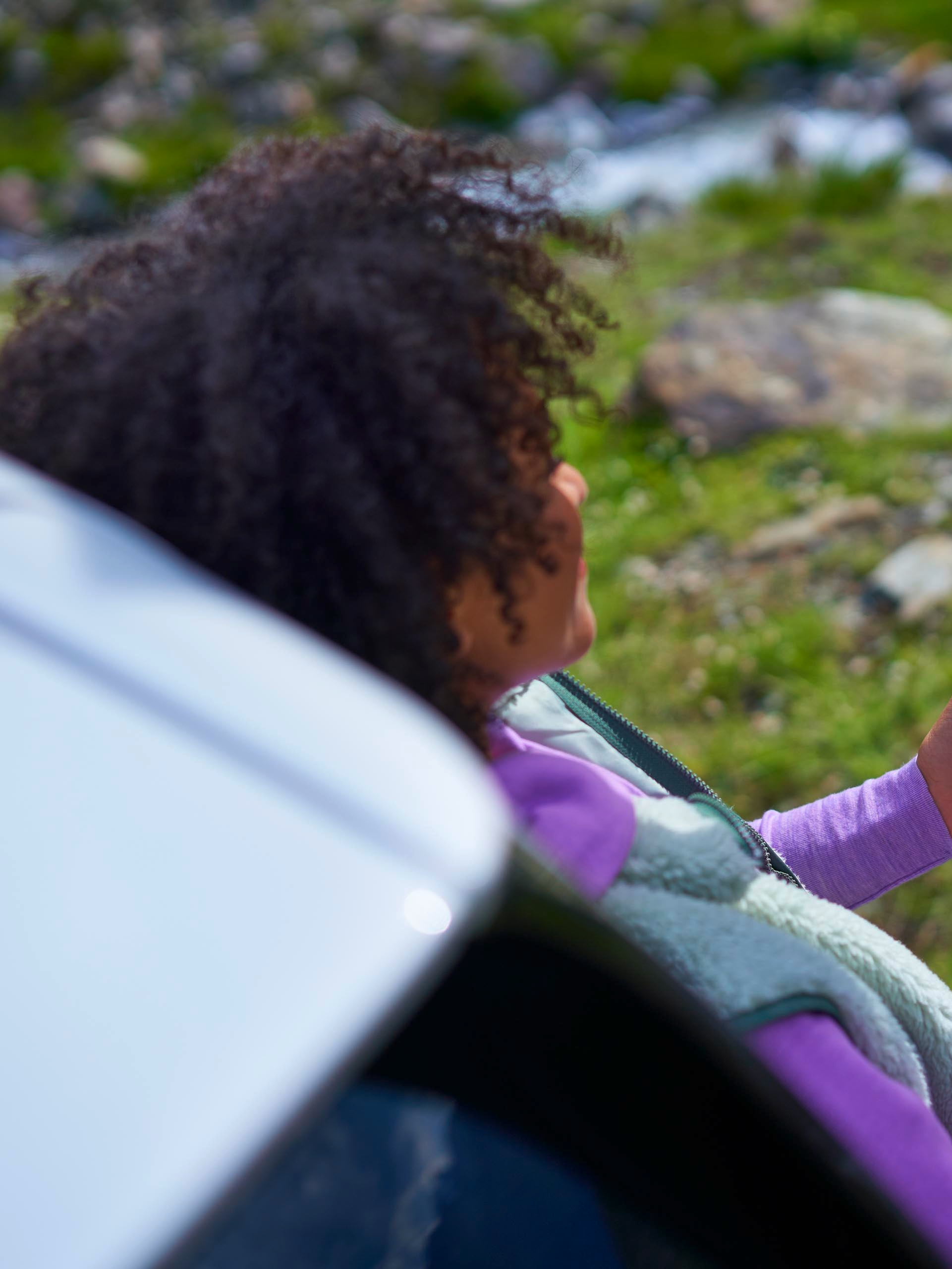 Oseba sedi zunaj poleg avtomobila in s pametnim telefonom preverja informacijo o polnilni postaji za električna vozila. V ozadju je naravna pokrajina s travo, kamni in vodo.
