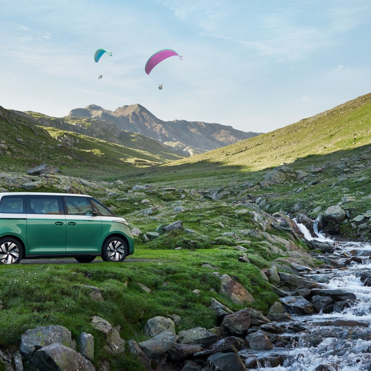 VW ID. Buzz steht an einem Bergbach mit geöffneter Heckklappe, zwei Wanderer laden Rucksäcke ein