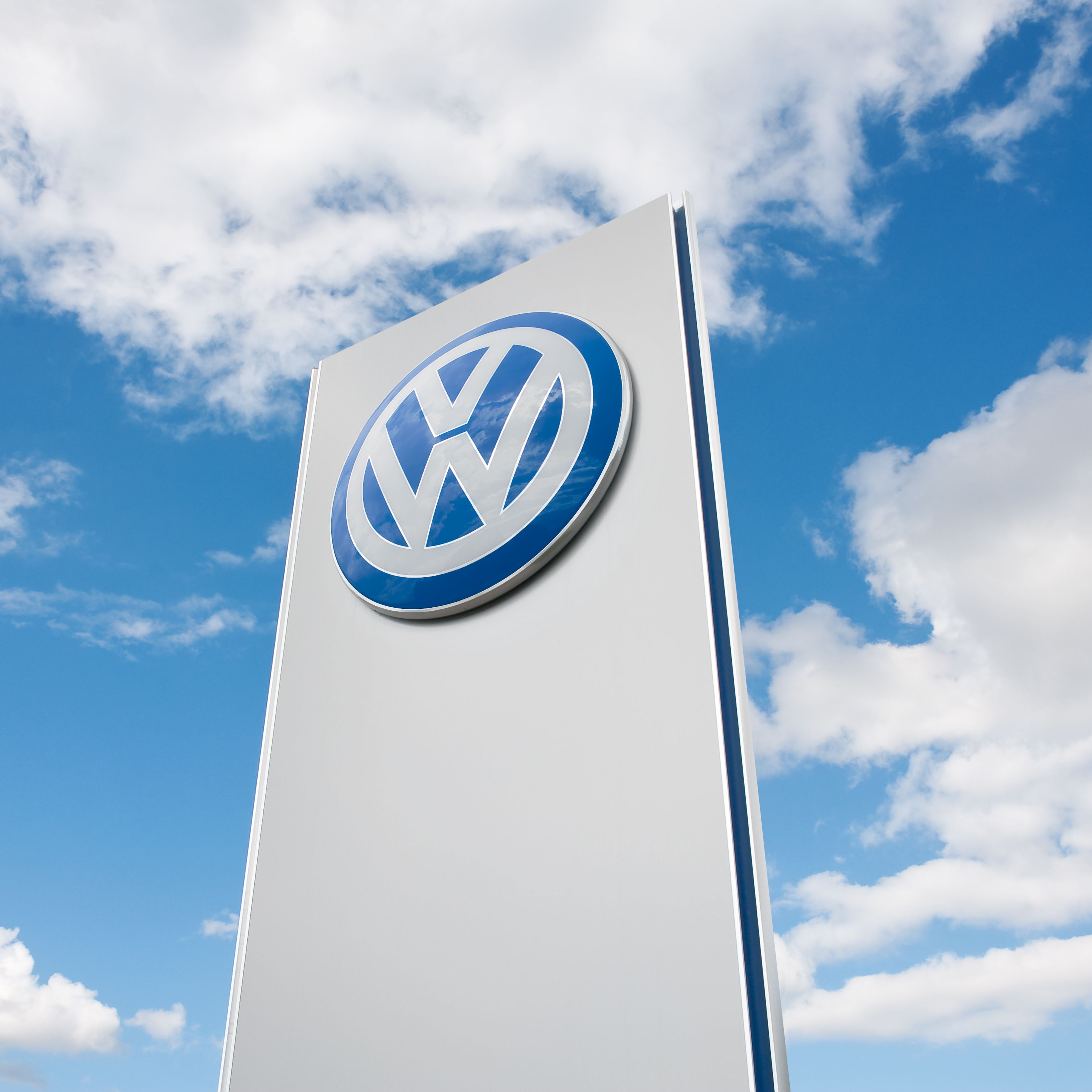 Na sliki je znak podjetja Volkswagen na visokem belem stebru, obdan z modrim nebom in belimi oblaki.