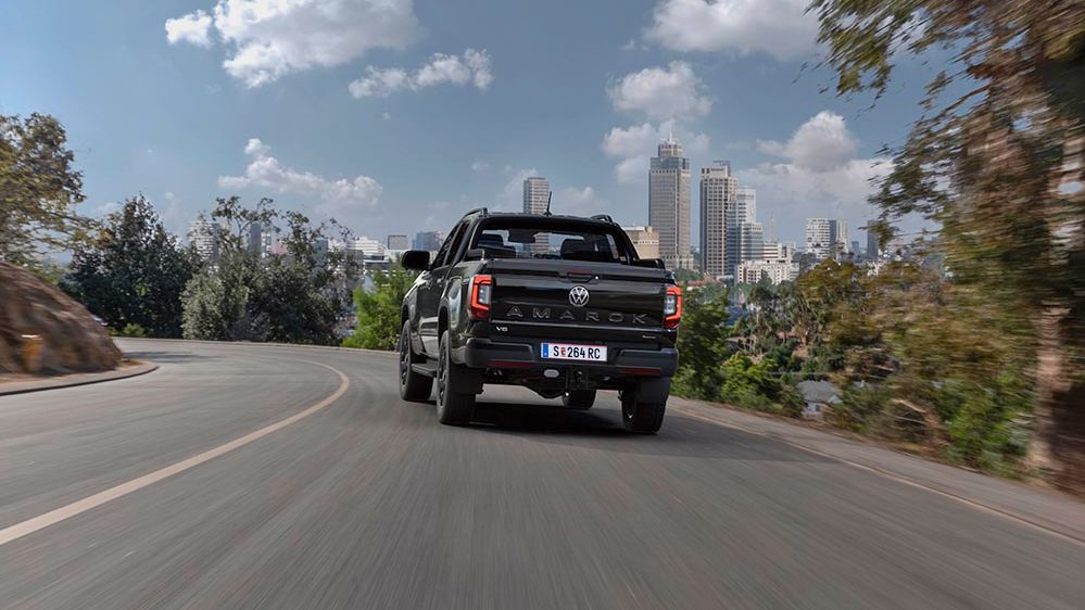 Ansicht eines schwarzen Pick-up-Trucks auf einer kurvigen Straße, während er zwischen Bäumen und moderner städtischer Skyline im Hintergrund fährt. Der Himmel ist blau mit einigen Wolken.