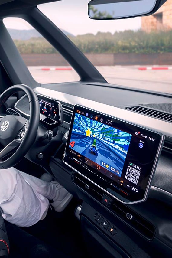 Ženska sedi na voznikovem sedežu avtomobila, gleda in igra video igro na velikem zaslonu infotainment sistema. V ozadju so vidne gore.