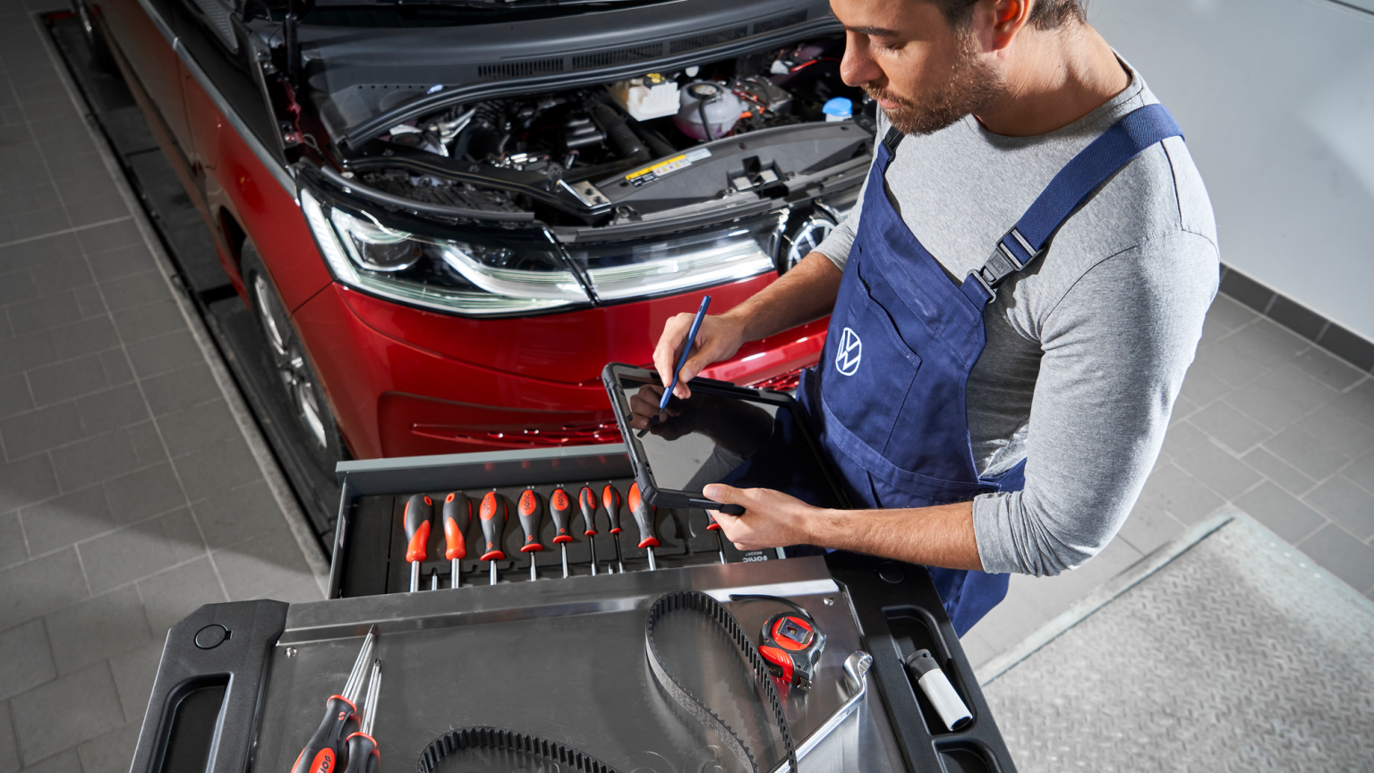 Avtomehanik v modrem kombinezonu stoji ob rdečem avtomobilu z odprtim pokrovom motorja in na tablični napravi vnaša podatke. Pred njim je voziček z orodjem, na katerem so izvijači, merilni kabel in druga delavniška oprema. Scena prikazuje servisni pregled ali popravilo vozila v delavnici.