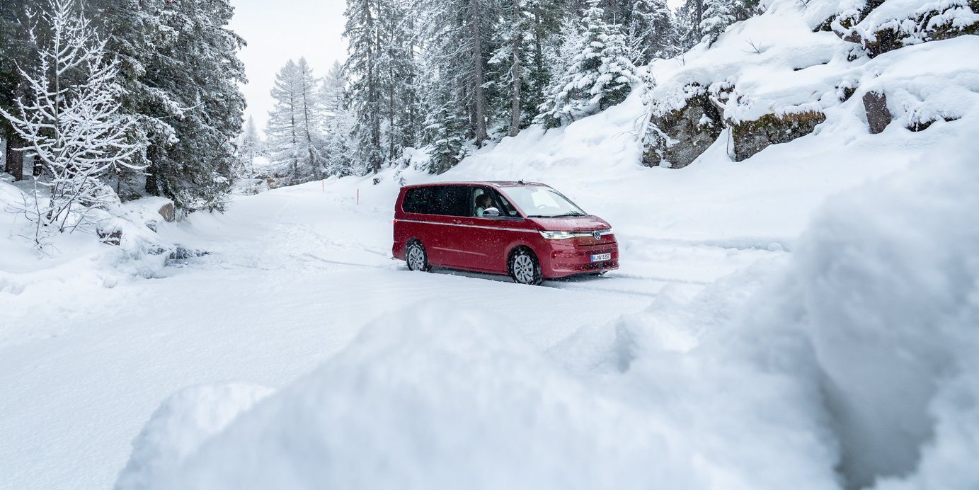Rdeč avtomobil vozi po zasneženi cesti, obdan z zasneženimi drevesi in skalami. Nebo je delno oblačno. Pokrajina je zimska in mirna.