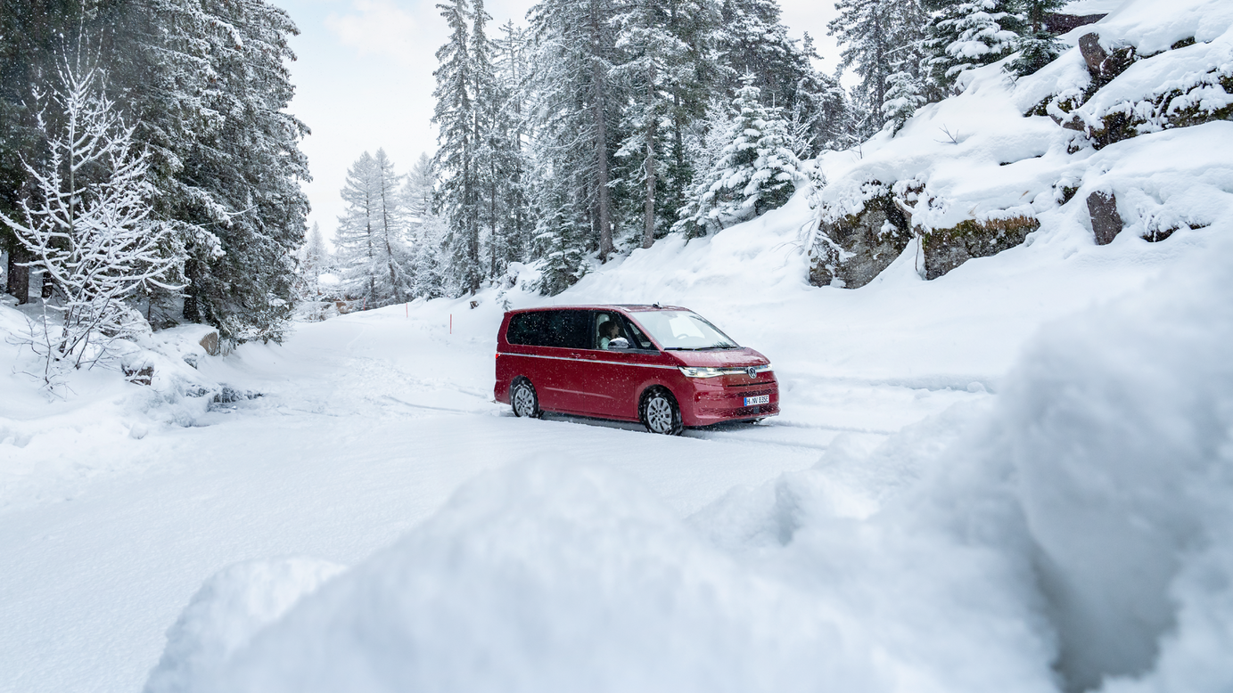 Rdeč avtomobil vozi po zasneženi cesti, obdan z zasneženimi drevesi in skalami. Nebo je delno oblačno. Pokrajina je zimska in mirna.