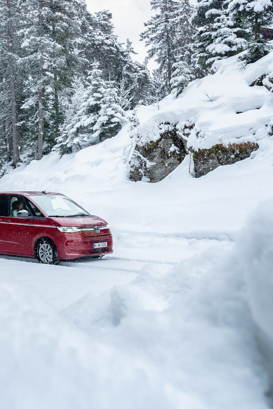 Rdeč avtomobil vozi po zasneženi cesti, obdan z zasneženimi drevesi in skalami. Nebo je delno oblačno. Pokrajina je zimska in mirna.