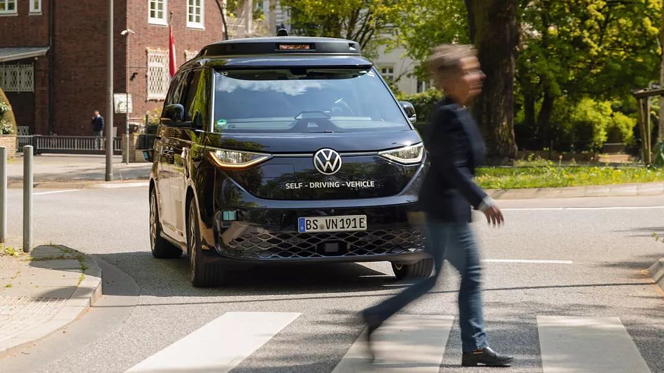 Ein selbstfahrendes Fahrzeug von Volkswagen fährt auf einer Straße. Eine Person überquert gerade den Zebrastreifen vor dem Auto. Im Hintergrund sind Bäume und Gebäude zu sehen.