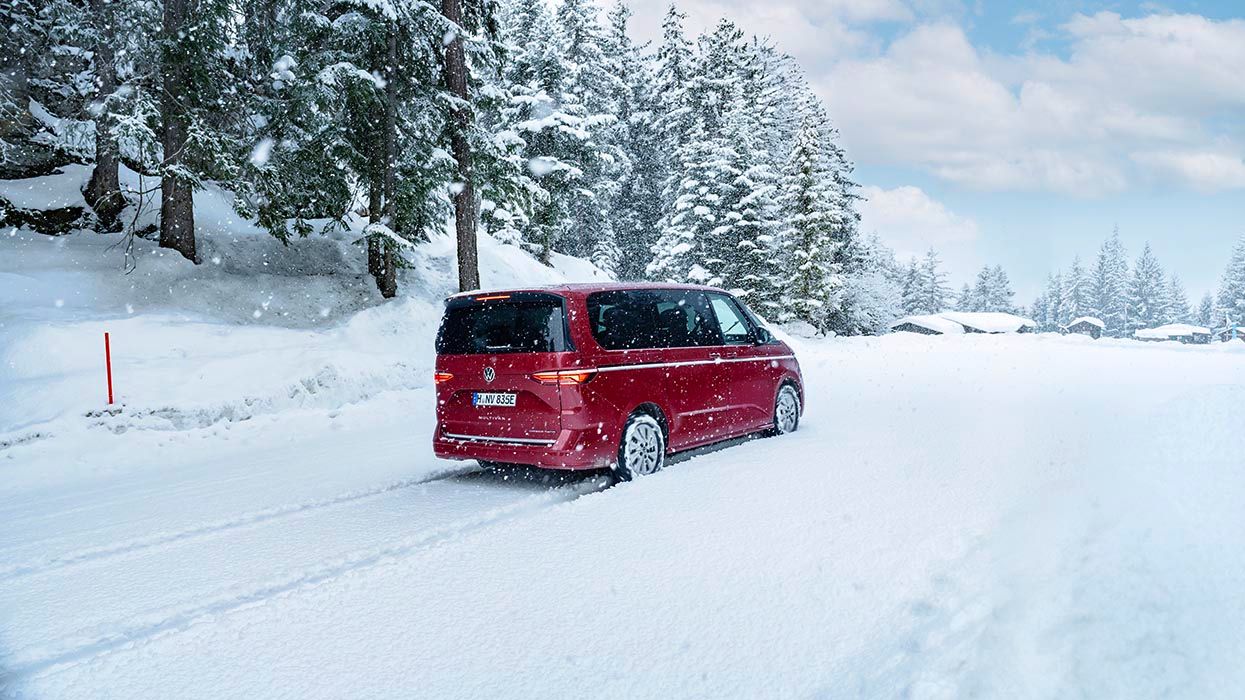 Rdeč minivan vozi po zasneženi cesti, obdani s snežno pokritimi drevesi. Nebo je oblačno, kar prispeva k zimskem vzdušju. Avto pušča sledi na snežni površini.