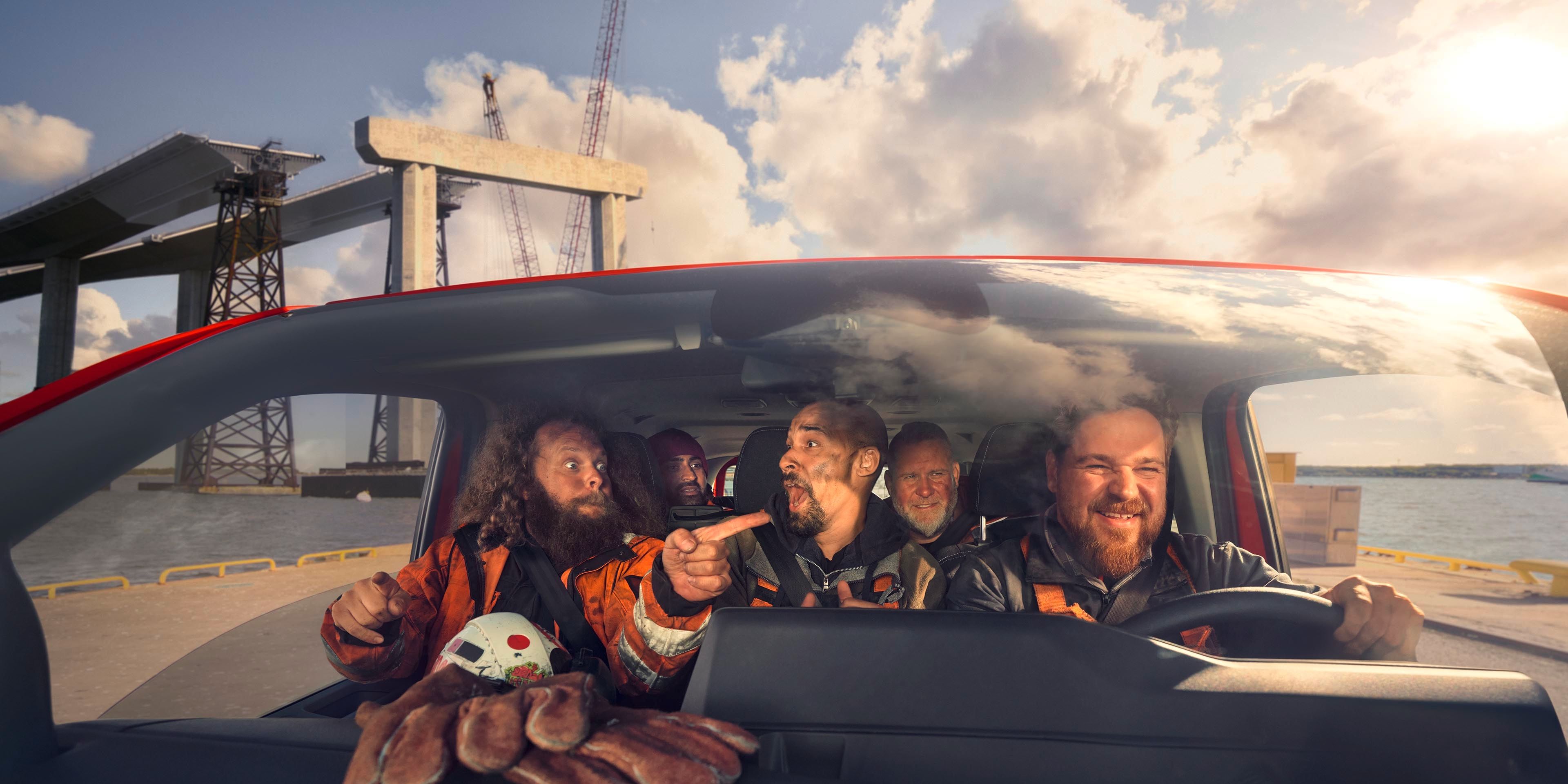 Skupina ljudi sedi v notranjosti vozila in gleda skozi sprednje vetrobransko steklo. Vozilo je parkirano ob vodi, v ozadju so vidni gradbeni žerjavi. Na armaturni plošči je plišasta igrača.