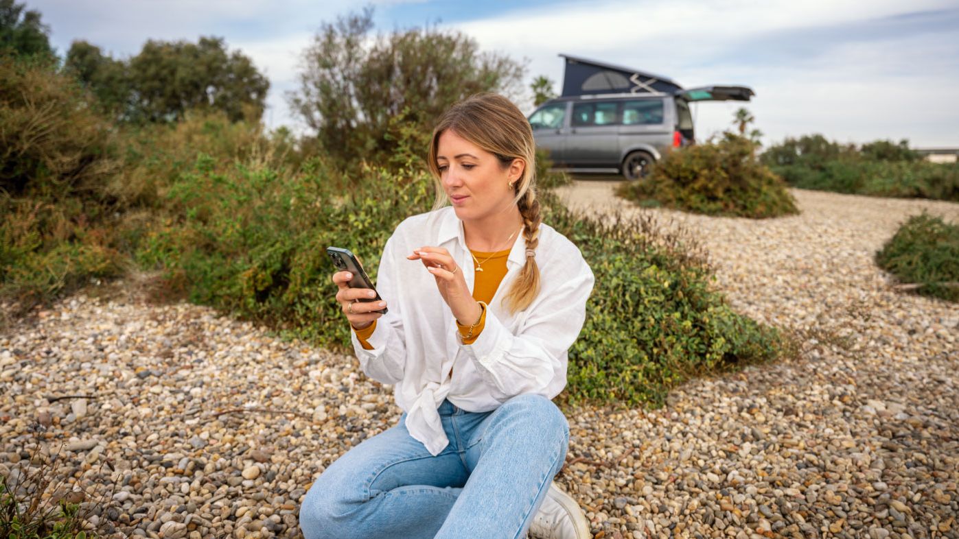 Eine Frau sitzt im Freien am Boden und blickt auf ihr Smartphone, im Hintergrund ein VW California