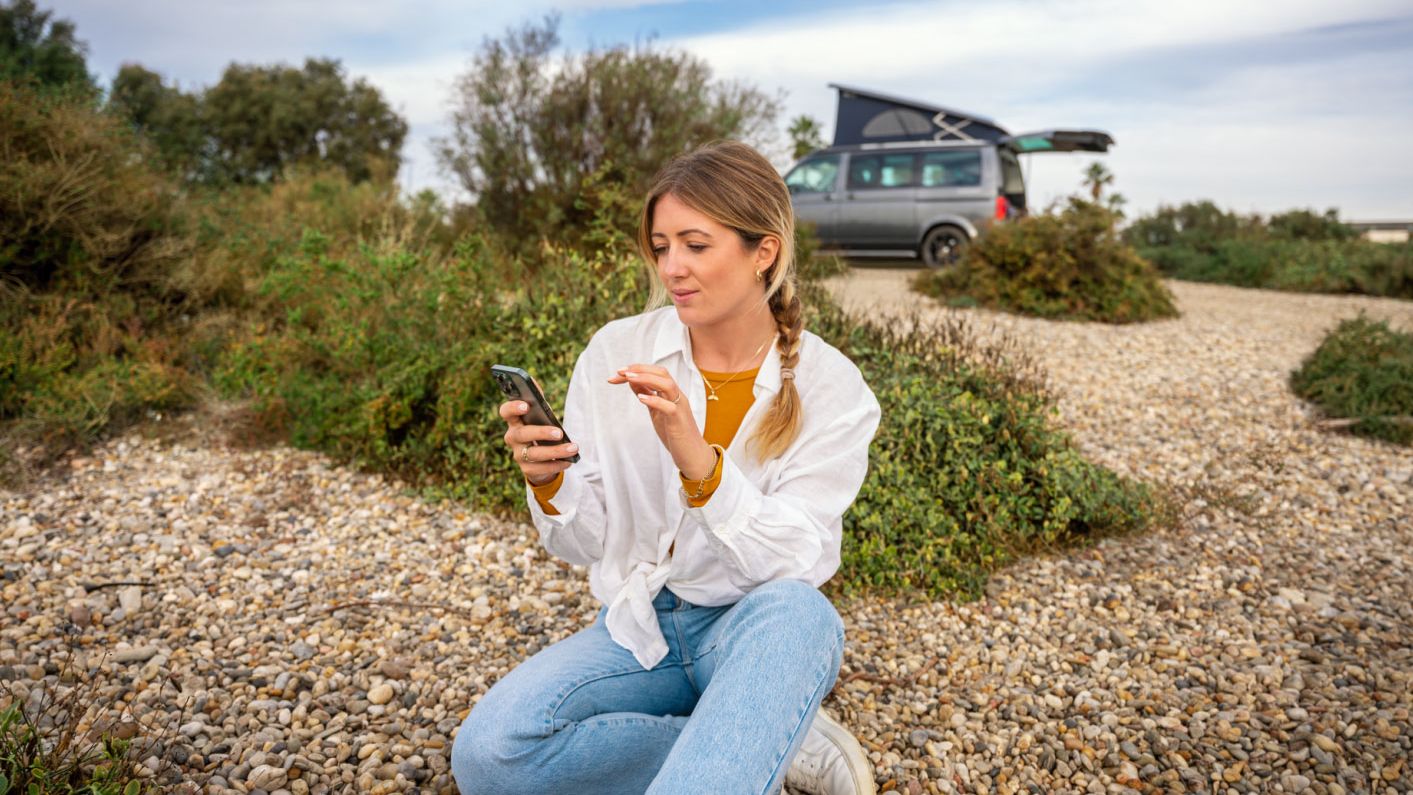 Eine Frau sitzt im Freien am Boden und blickt auf ihr Smartphone, im Hintergrund ein VW California