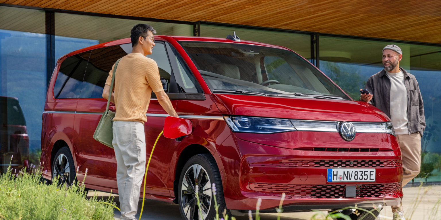 Dva moška stojita poleg rdečega Volkswagnovega kombija in se pripravljata na polnjenje vozila z elektriko. Eden drži kabel za polnjenje, drugi ima pametni telefon v roki. V ozadju je moderna steklena zgradba.