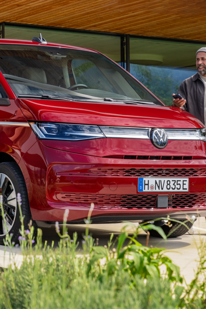 Dva moška stojita poleg rdečega Volkswagnovega kombija in se pripravljata na polnjenje vozila z elektriko. Eden drži kabel za polnjenje, drugi ima pametni telefon v roki. V ozadju je moderna steklena zgradba.