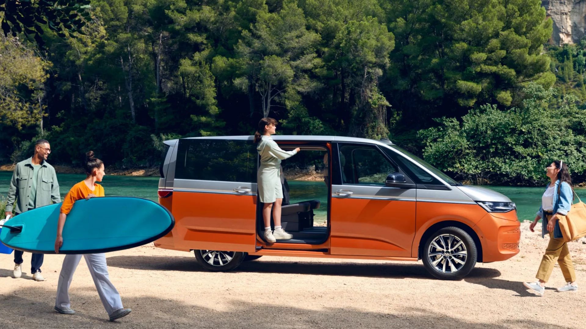 Seitenansicht eines VW Multivan mit geöffneter Schiebetüre an einem Fluss, eine Familie bewegt sich um das Auto