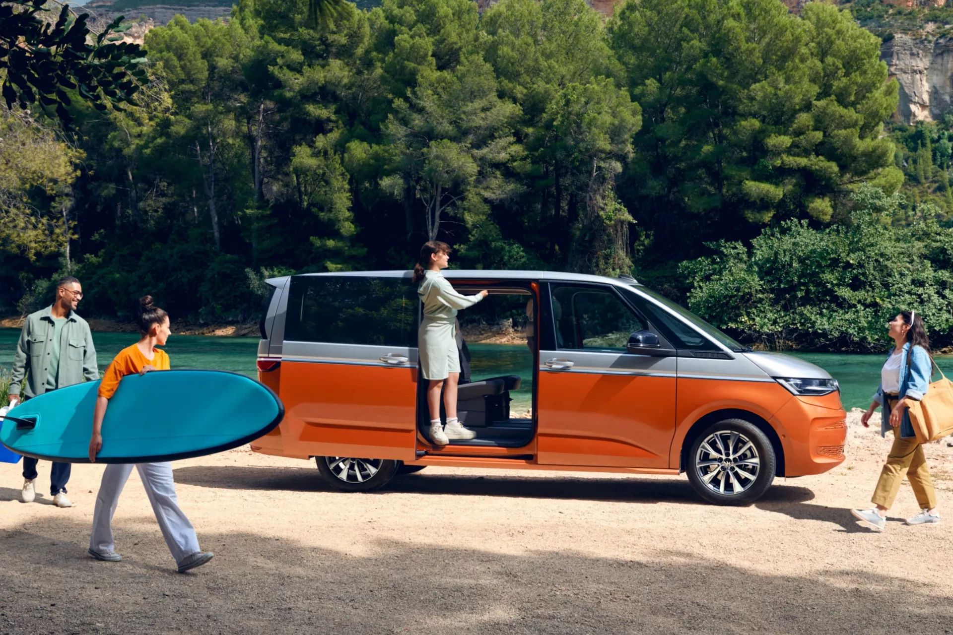 Seitenansicht eines VW Multivan mit geöffneter Schiebetüre an einem Fluss, eine Familie bewegt sich um das Auto