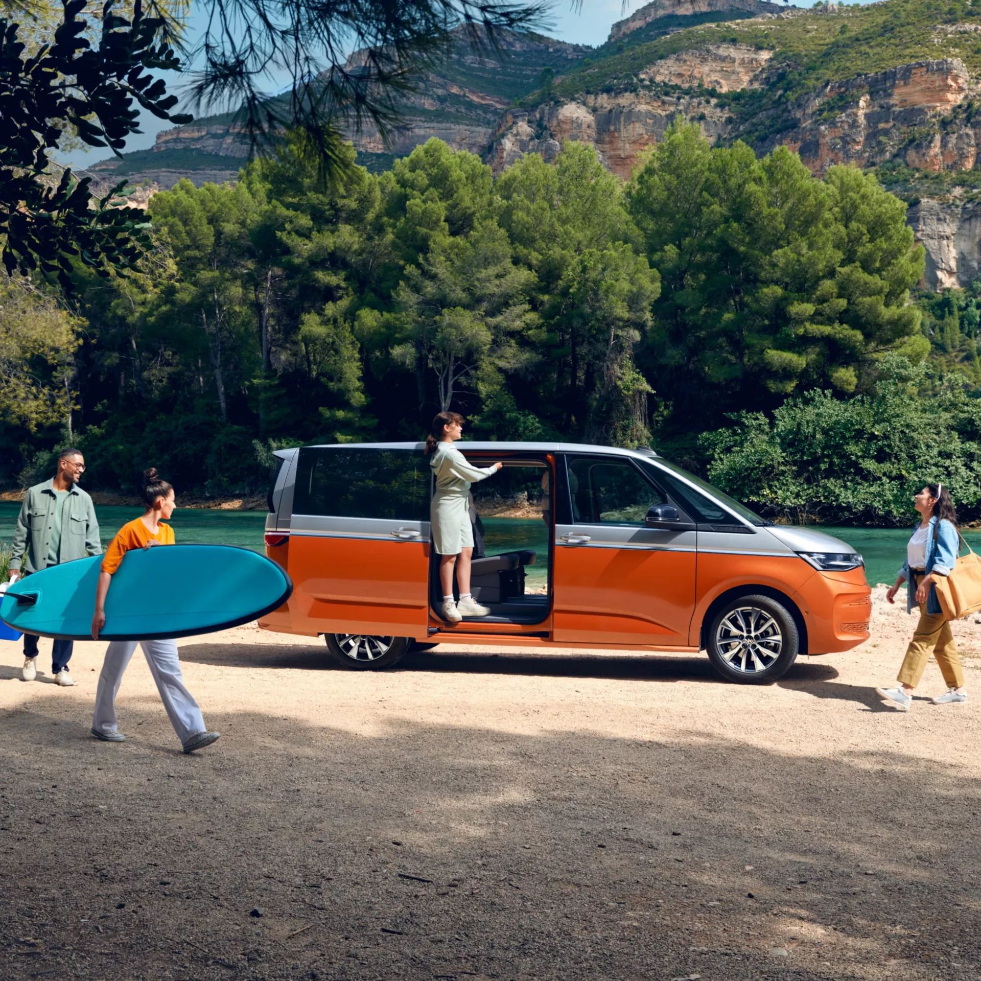 Seitenansicht eines VW Multivan mit geöffneter Schiebetüre an einem Fluss, eine Familie bewegt sich um das Auto