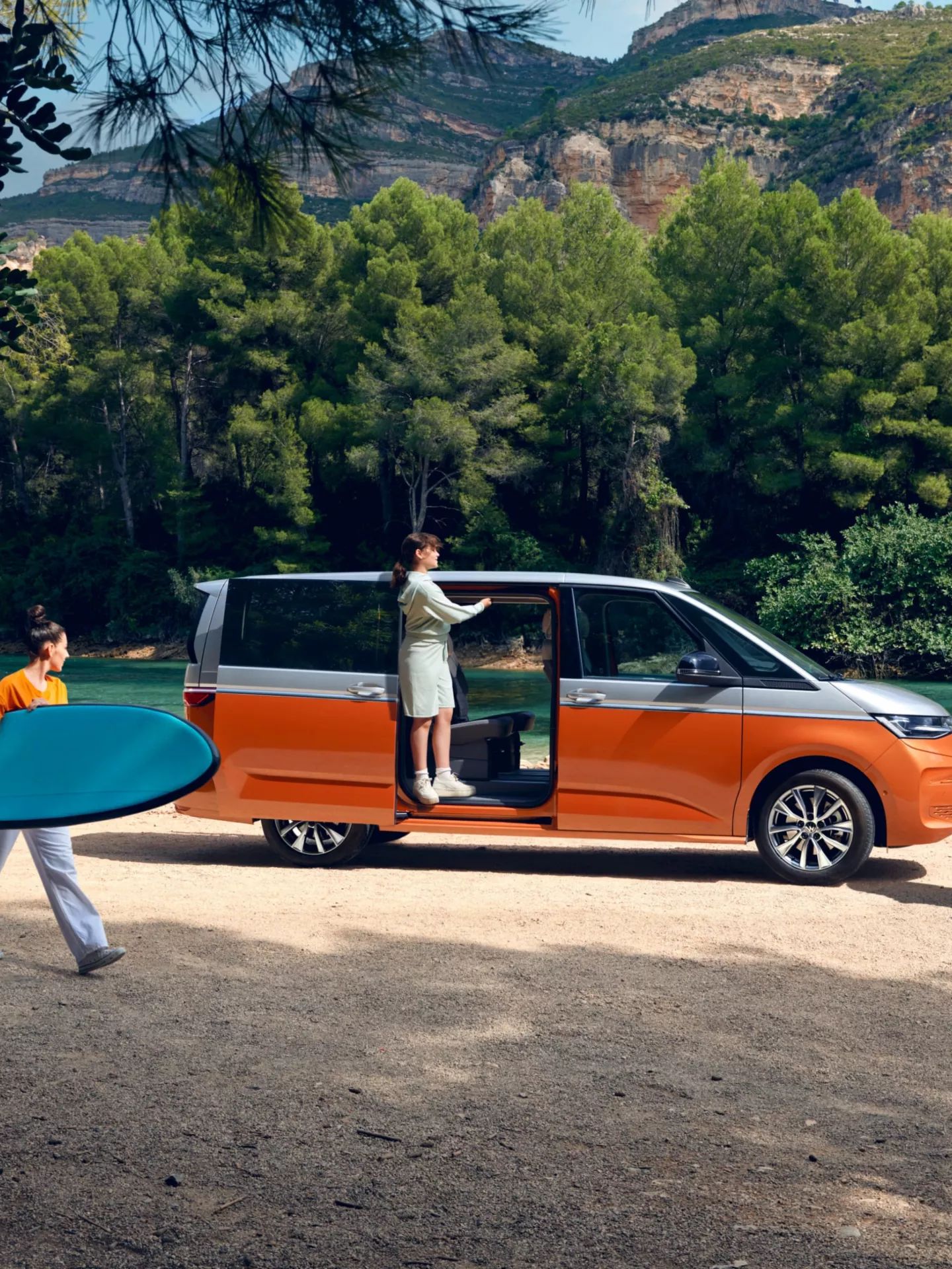 Seitenansicht eines VW Multivan mit geöffneter Schiebetüre an einem Fluss, eine Familie bewegt sich um das Auto