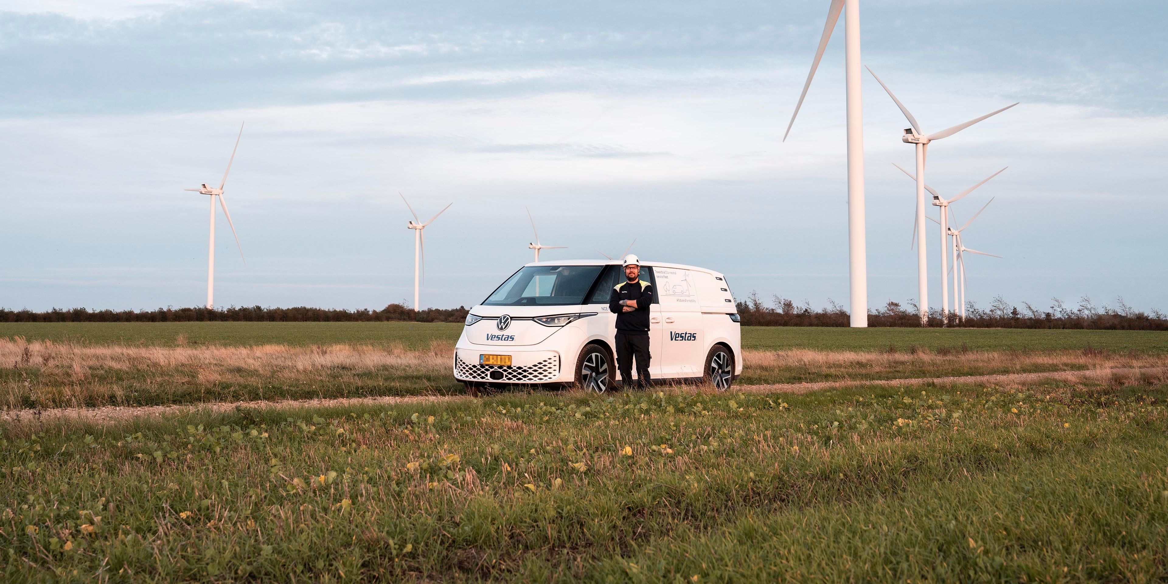 Bel kombi vozilo je parkirano na travniku v bližini več vetrnic. Oseba stoji zraven vozila. Na nebu so oblaki.