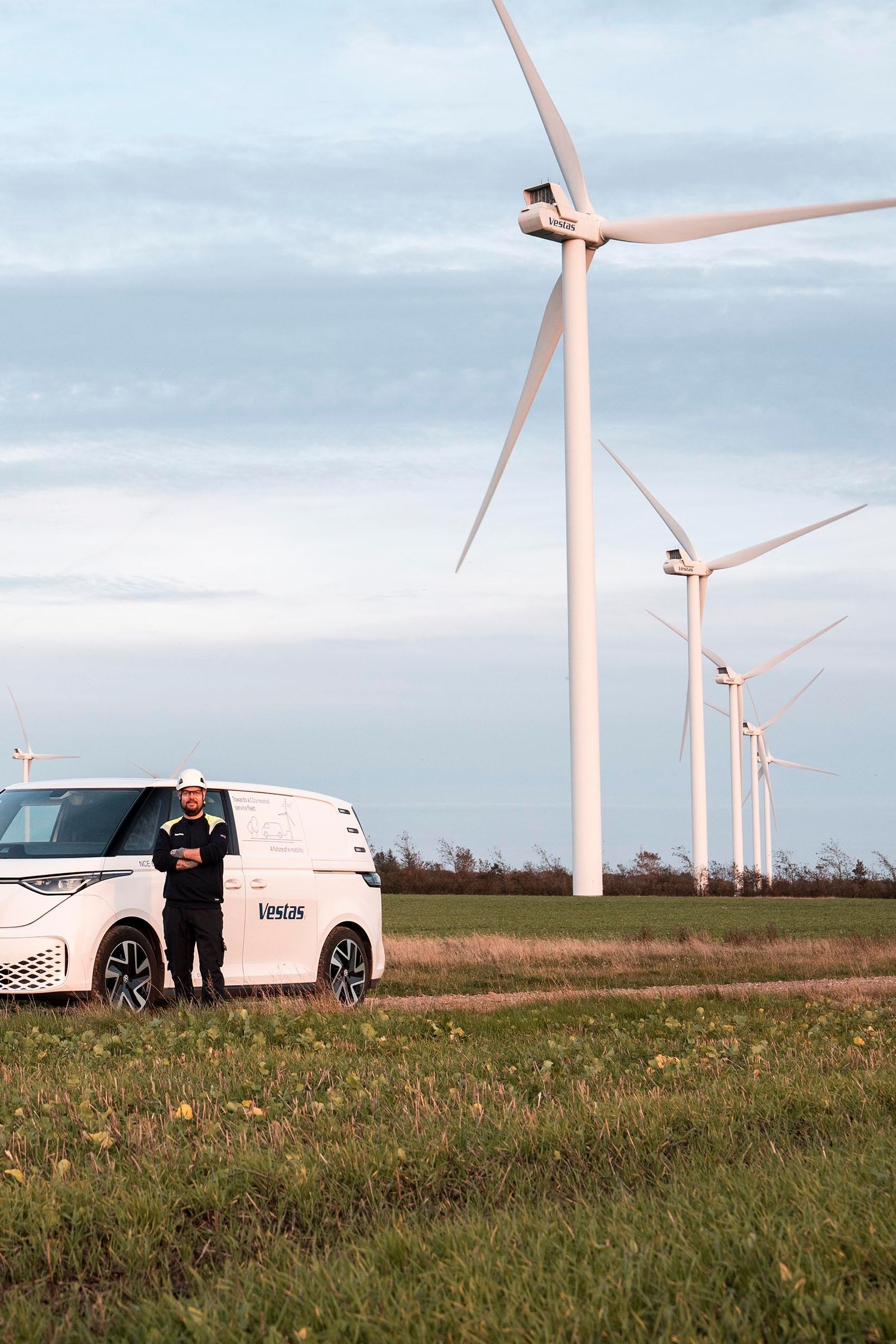 Bel kombi vozilo je parkirano na travniku v bližini več vetrnic. Oseba stoji zraven vozila. Na nebu so oblaki.