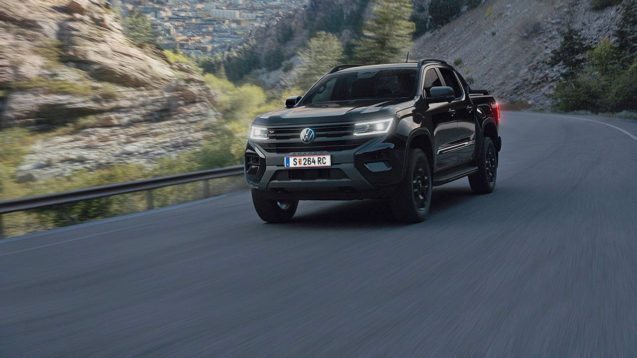 Ein schwarzer Pickup fährt entlang einer kurvenreichen Bergstraße. Im Hintergrund sind steile Felsen und eine Landschaft mit Bäumen und einer Stadt zu sehen. Der Himmel ist teilweise bewölkt.