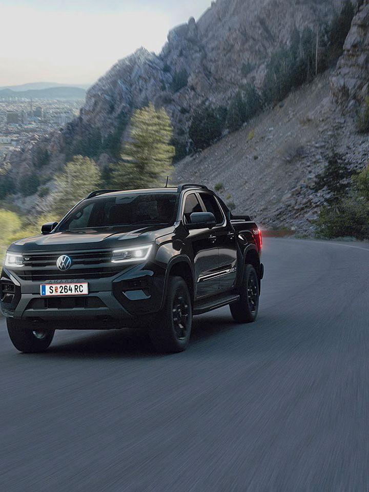 Ein schwarzer Pickup fährt entlang einer kurvenreichen Bergstraße. Im Hintergrund sind steile Felsen und eine Landschaft mit Bäumen und einer Stadt zu sehen. Der Himmel ist teilweise bewölkt.