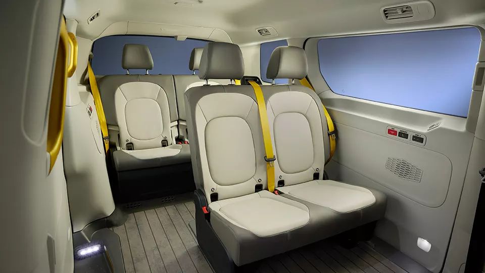 Innenraum eines modernen Fahrzeugs mit zwei Sitzreihen. Sitze sind hell und haben gelbe Sicherheitsgurte. Der Boden ist aus Holzpaneelen.